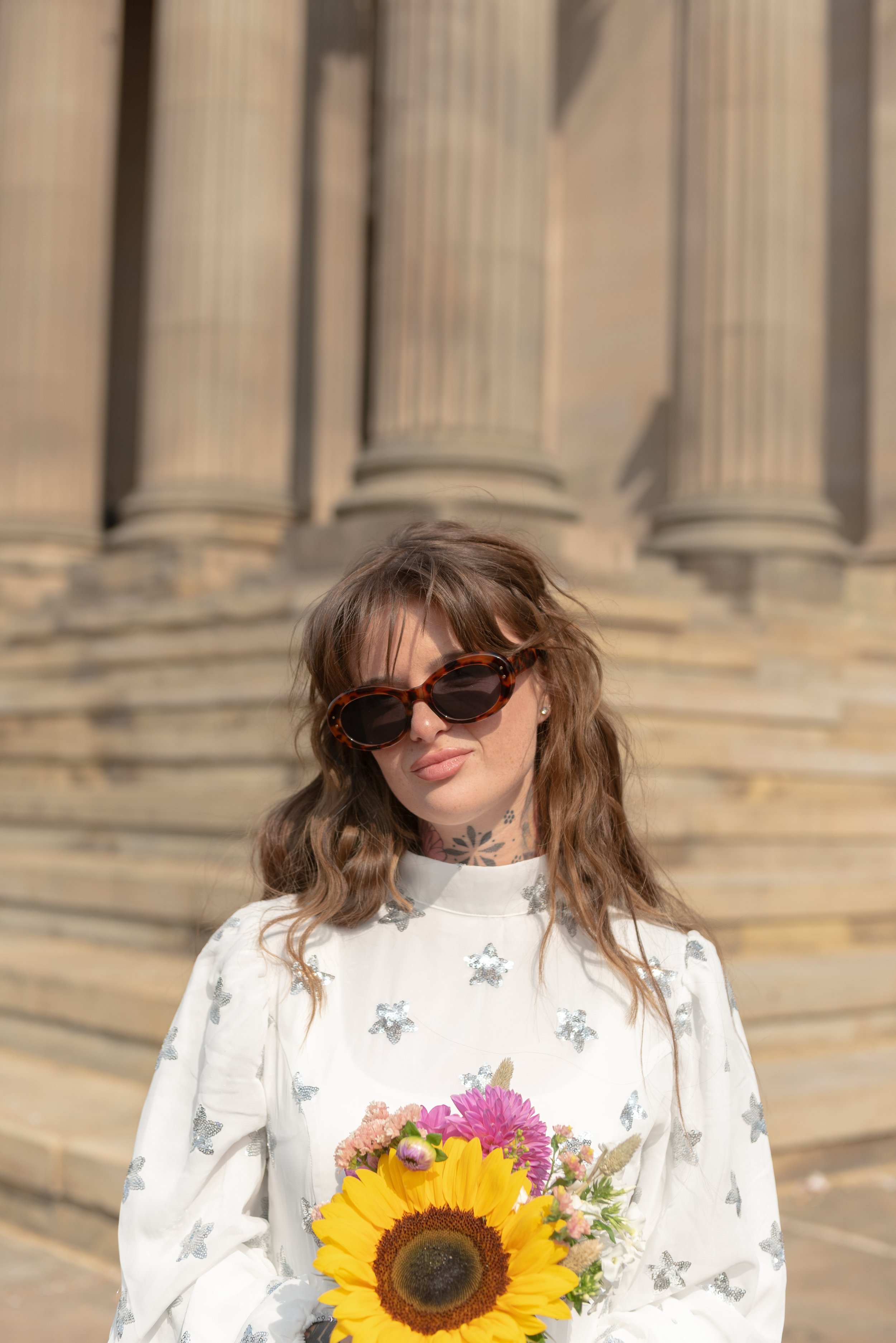Woman wearing large dark sunglasses and a white dress with silver butterflies, holding a bouquet of sunflowers and pink flowers, standing in front of a stone building with columns.