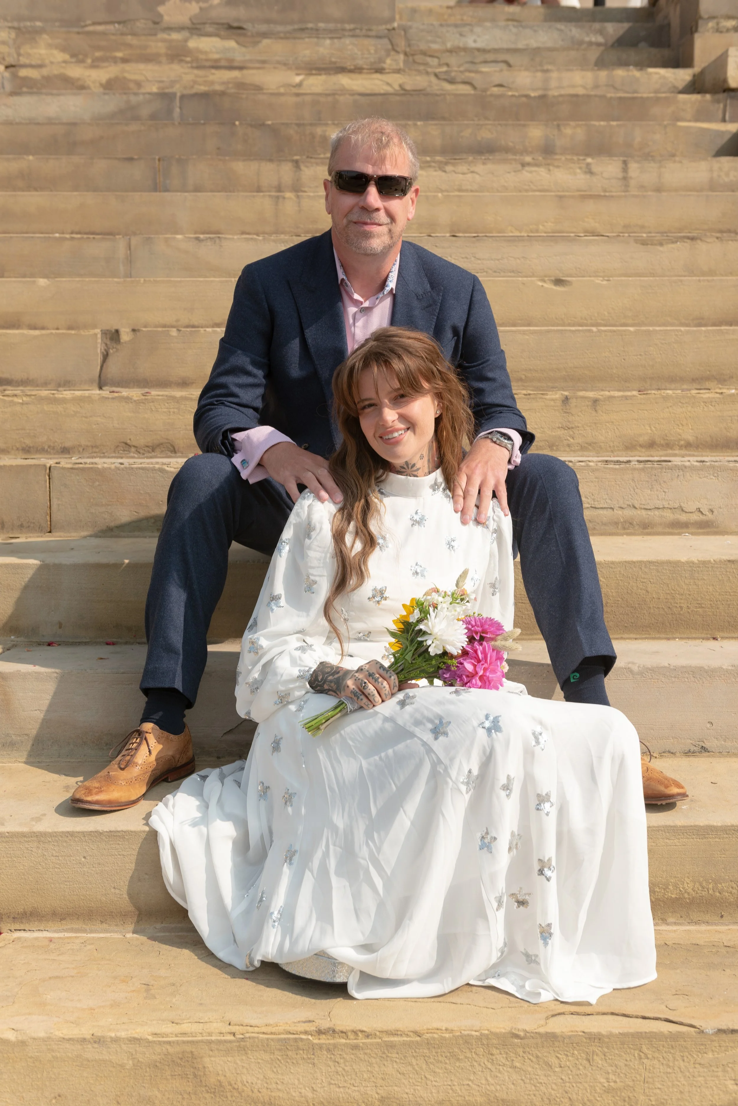 A man and woman sitting on stone steps outdoors, smiling. The woman in a white dress with butterfly embellishments holds a bouquet of flowers. The man wears a blue blazer, pink shirt, and sunglasses.
