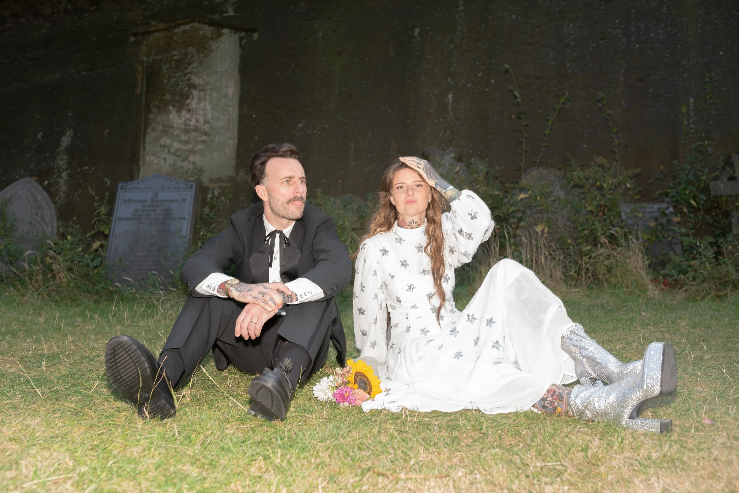 Man and woman sitting on grass at night, woman holding flowers, woman wearing silver boots, man wearing black suit, sitting near gravestones, outdoors with dark background and bushes.