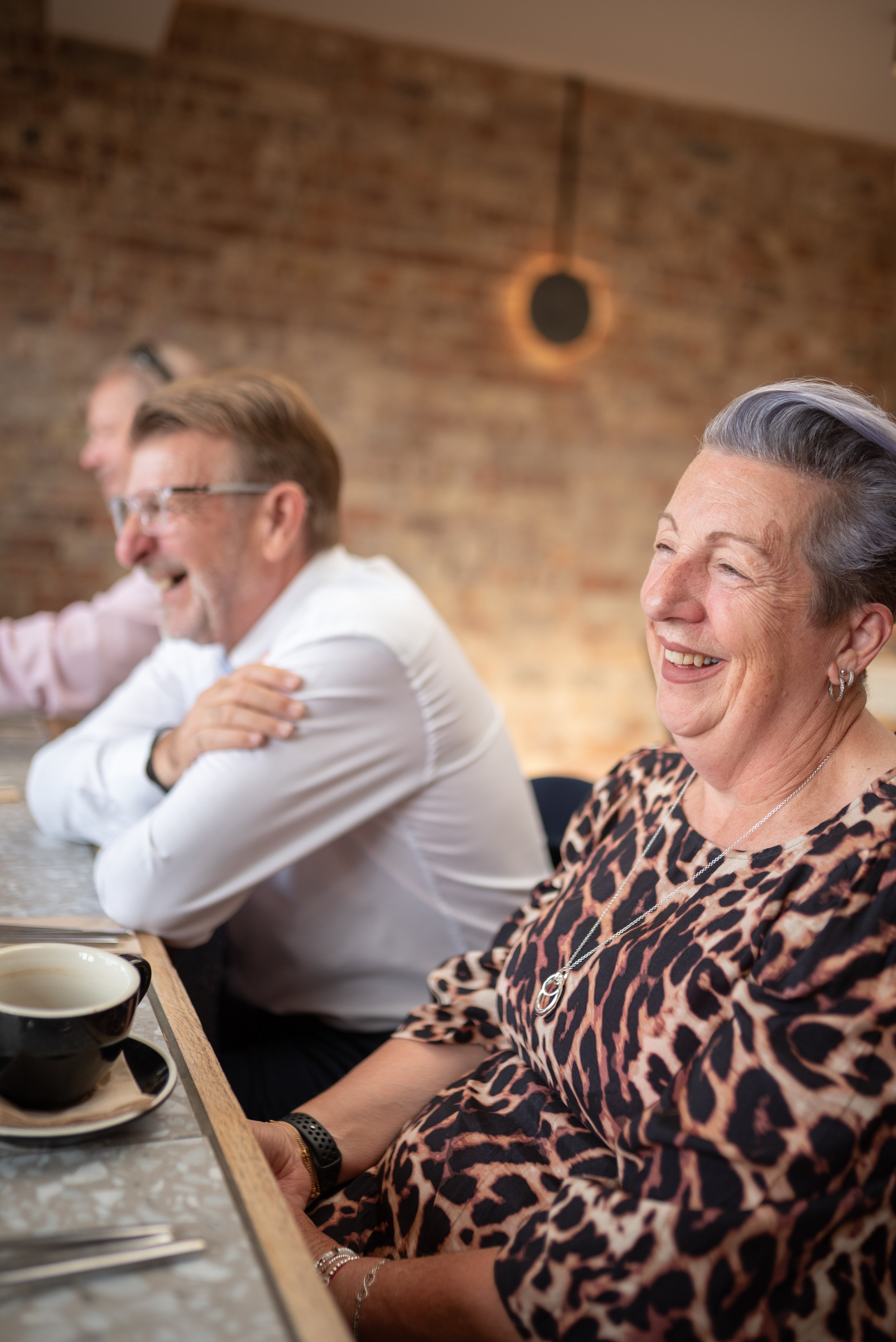 A group of senior adults sitting at a bar or cafe, smiling and laughing together, against a brick wall background.