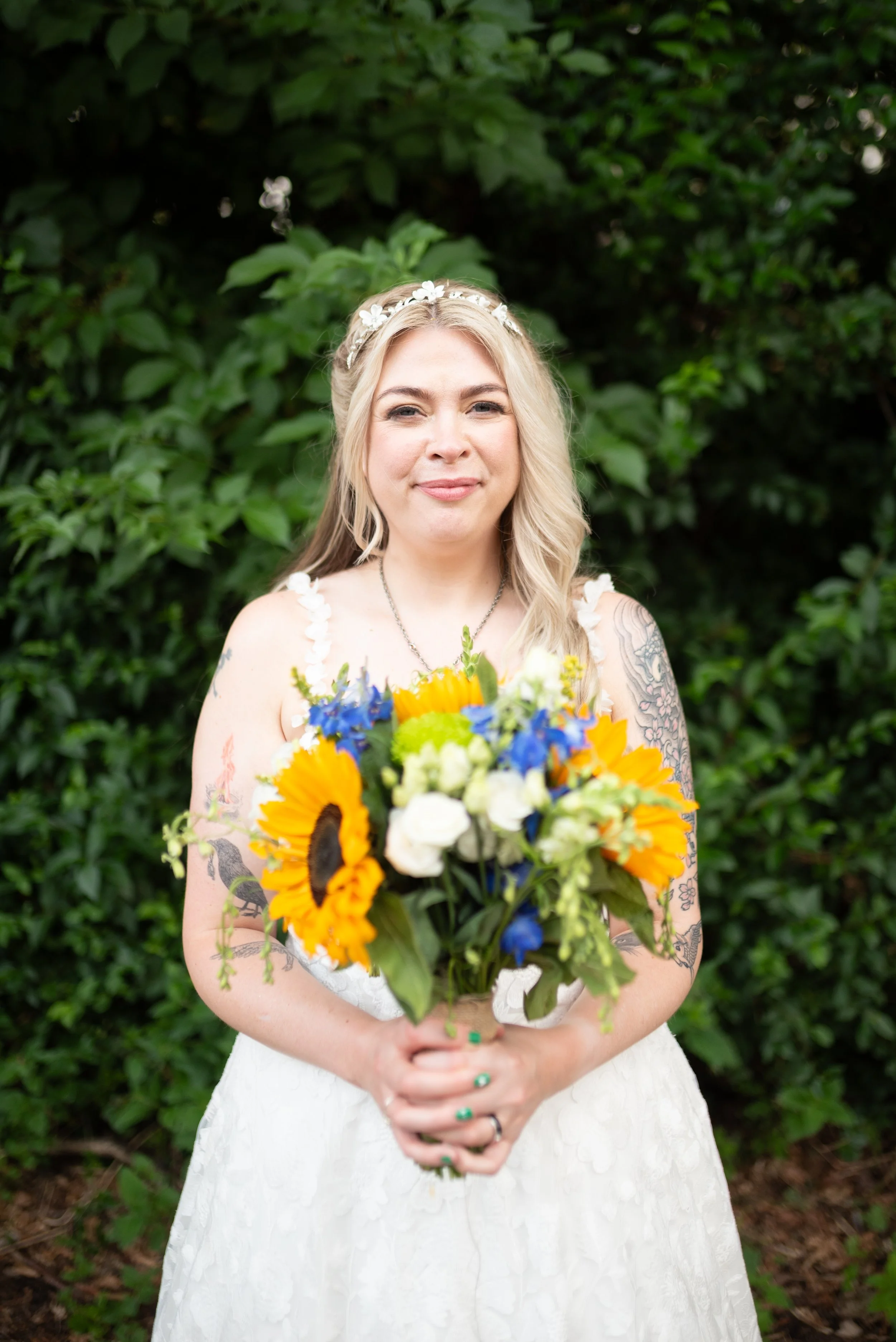 A woman in a wedding dress holding a bouquet of yellow sunflowers, white roses, and blue flowers, standing outdoors in front of green foliage.