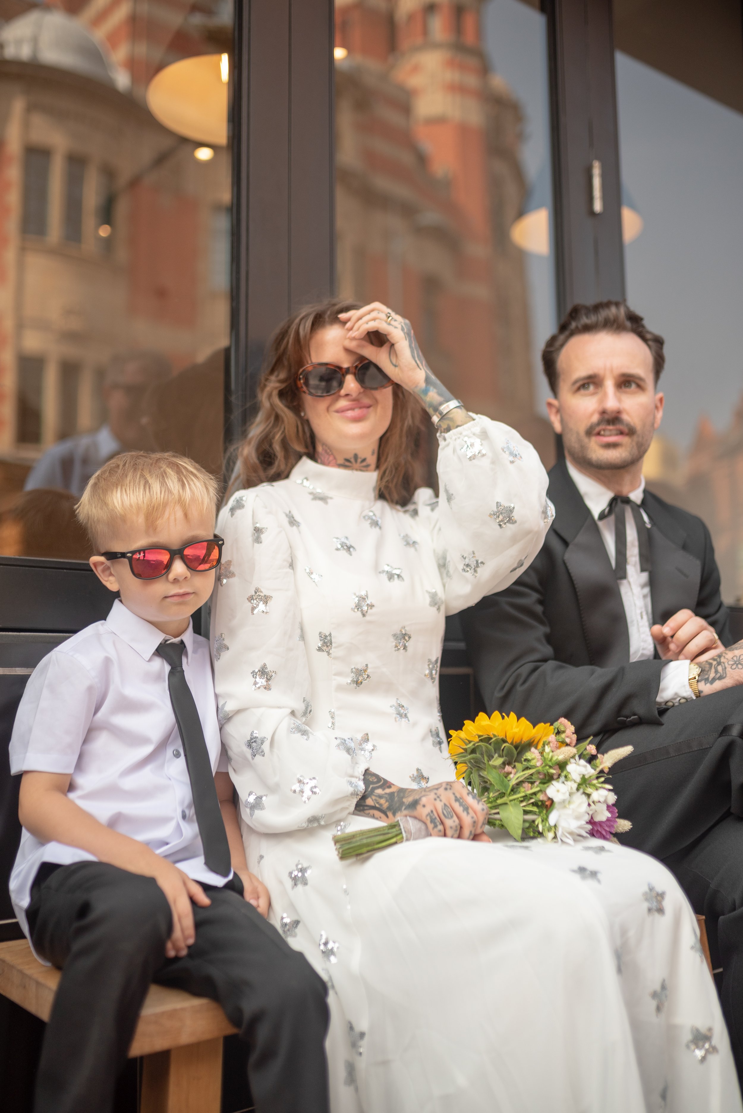 A woman, a young boy, and a man sitting outside in front of a building, with the woman holding a bouquet of flowers. The woman and boy are wearing sunglasses, and the woman is dressed in a white dress with silver butterfly decorations. The man is dre