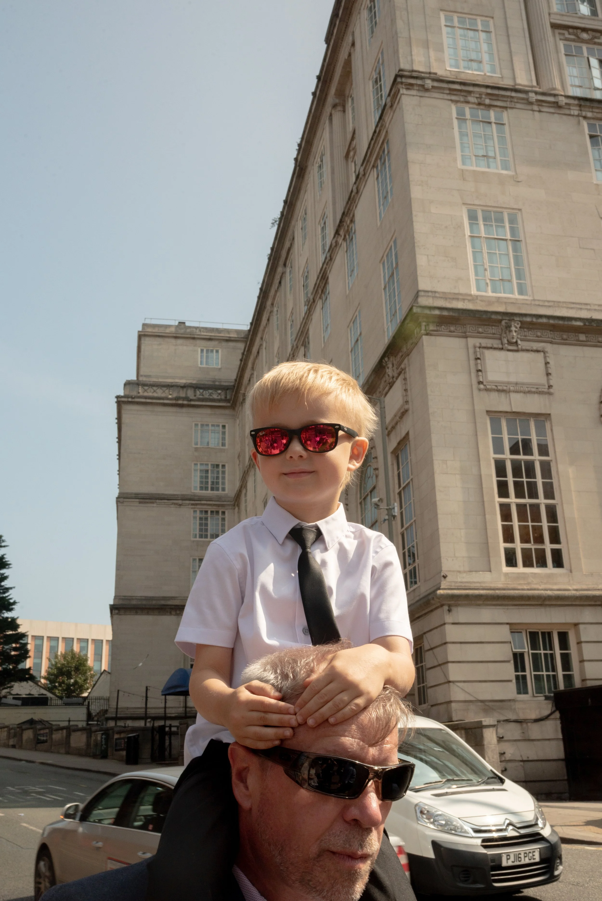 A young boy with blond hair, wearing a white shirt, black tie, and reflective sunglasses, sitting on an older man's shoulders. The older man has gray hair, a beard, and is also wearing sunglasses. They are outdoors in front of a large stone building 