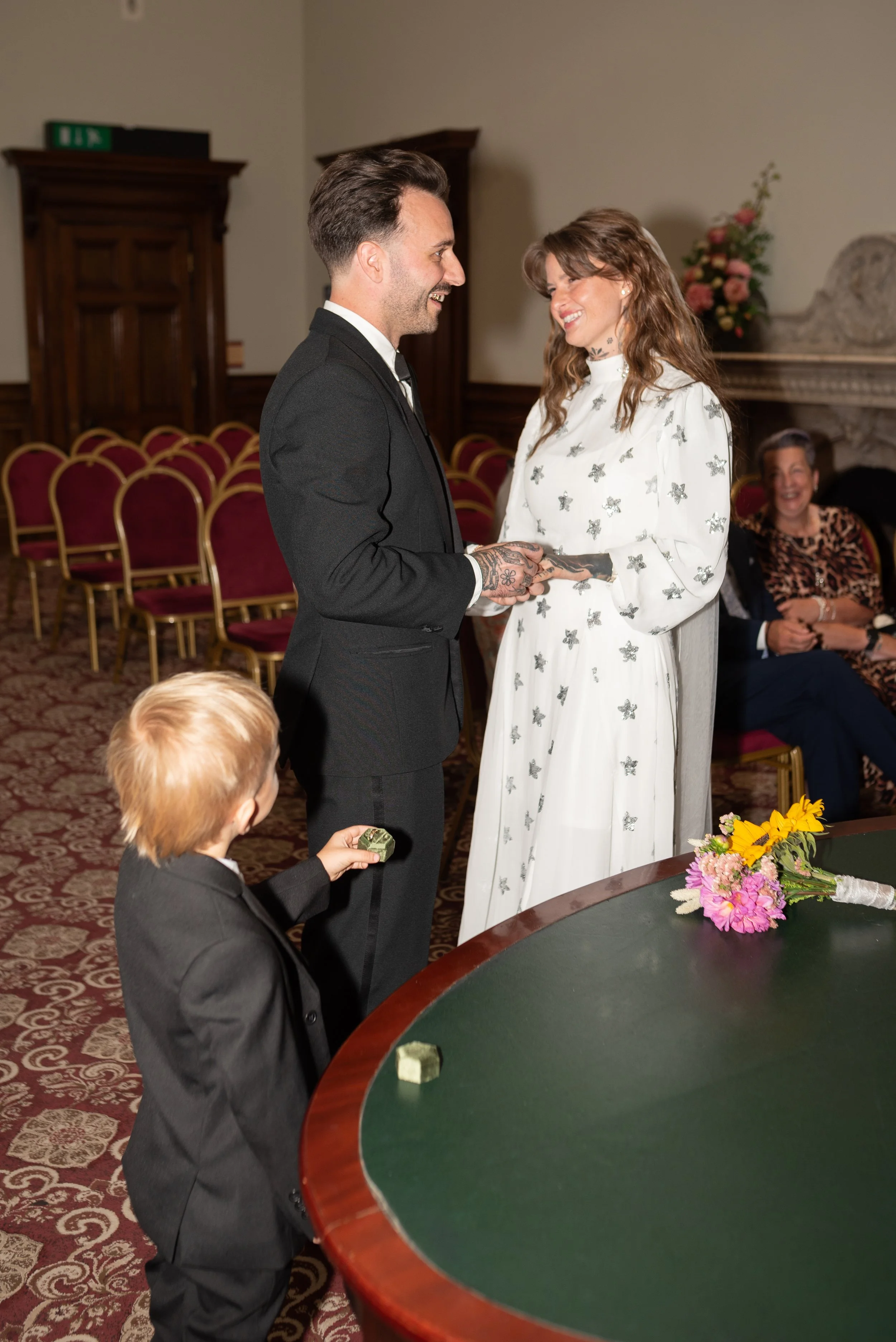 A couple getting married, holding hands and smiling at each other during the ceremony in a room with red chairs, a fireplace, and flowers on a table. A young boy in a black suit is holding a piece of cake, watching the couple.