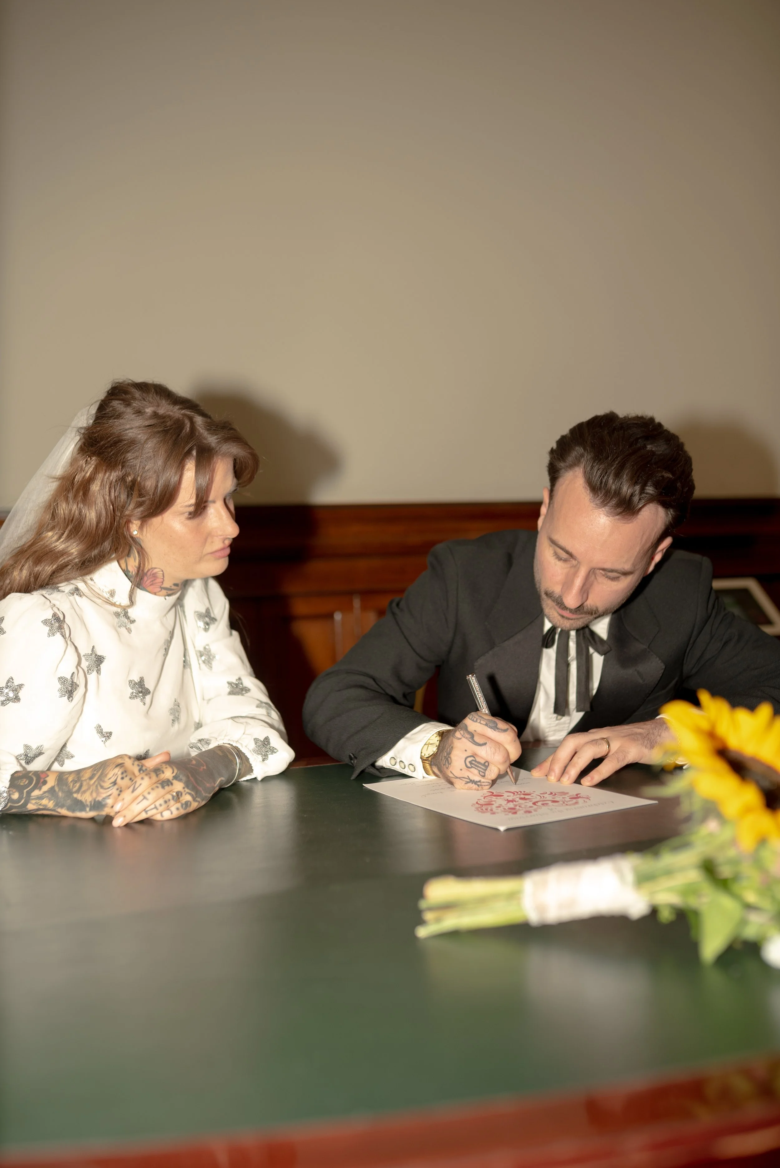 A couple getting married, with the woman wearing a white dress with butterfly patterns and a veil, and the man in a dark suit, signing a document on a dark table with a bouquet of sunflowers in the foreground.