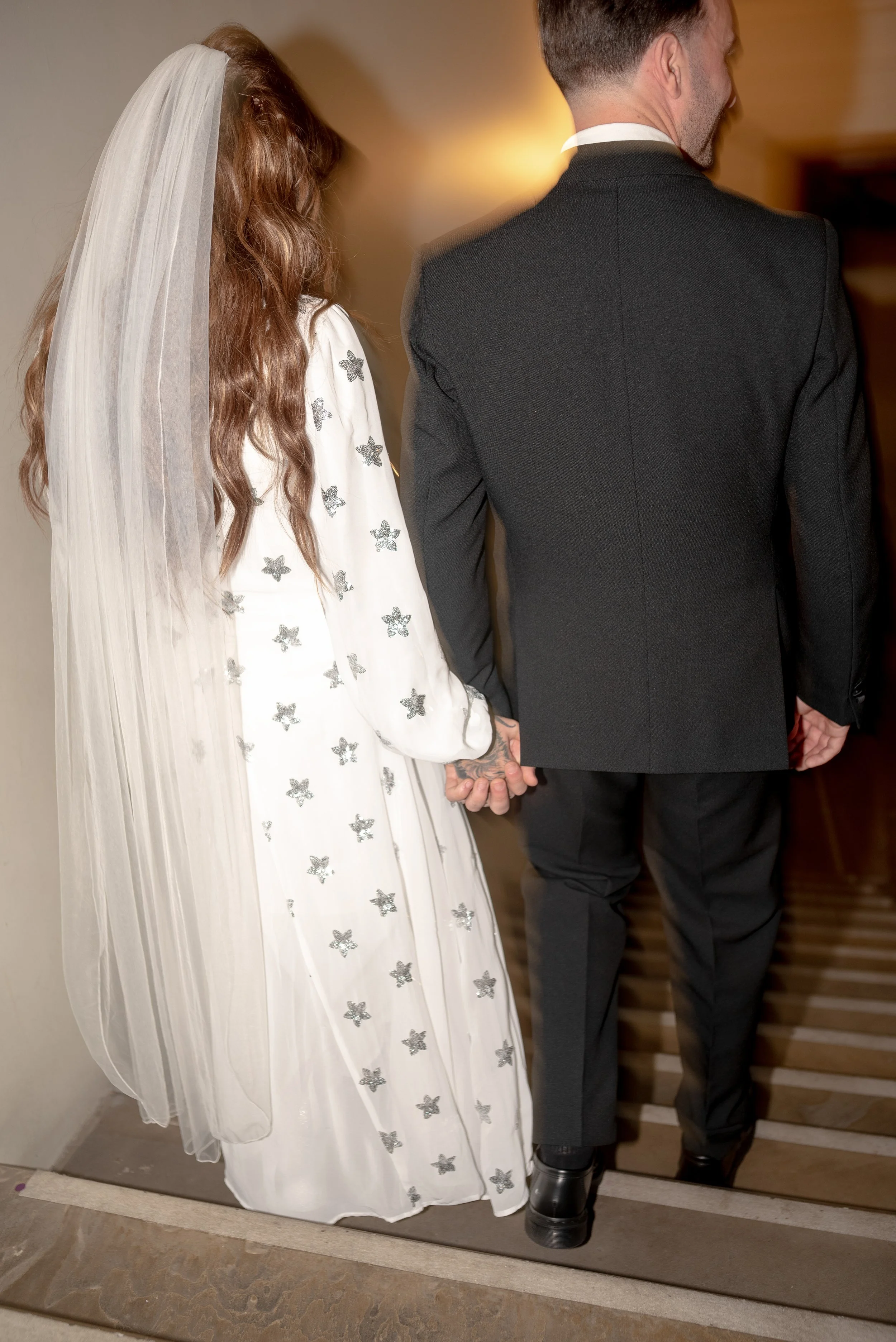 A bride and groom holding hands, with the bride's hand tattoo visible, walking away in wedding attire; the bride in a white gown with silver star embellishments and a veil, and the groom in a black suit.