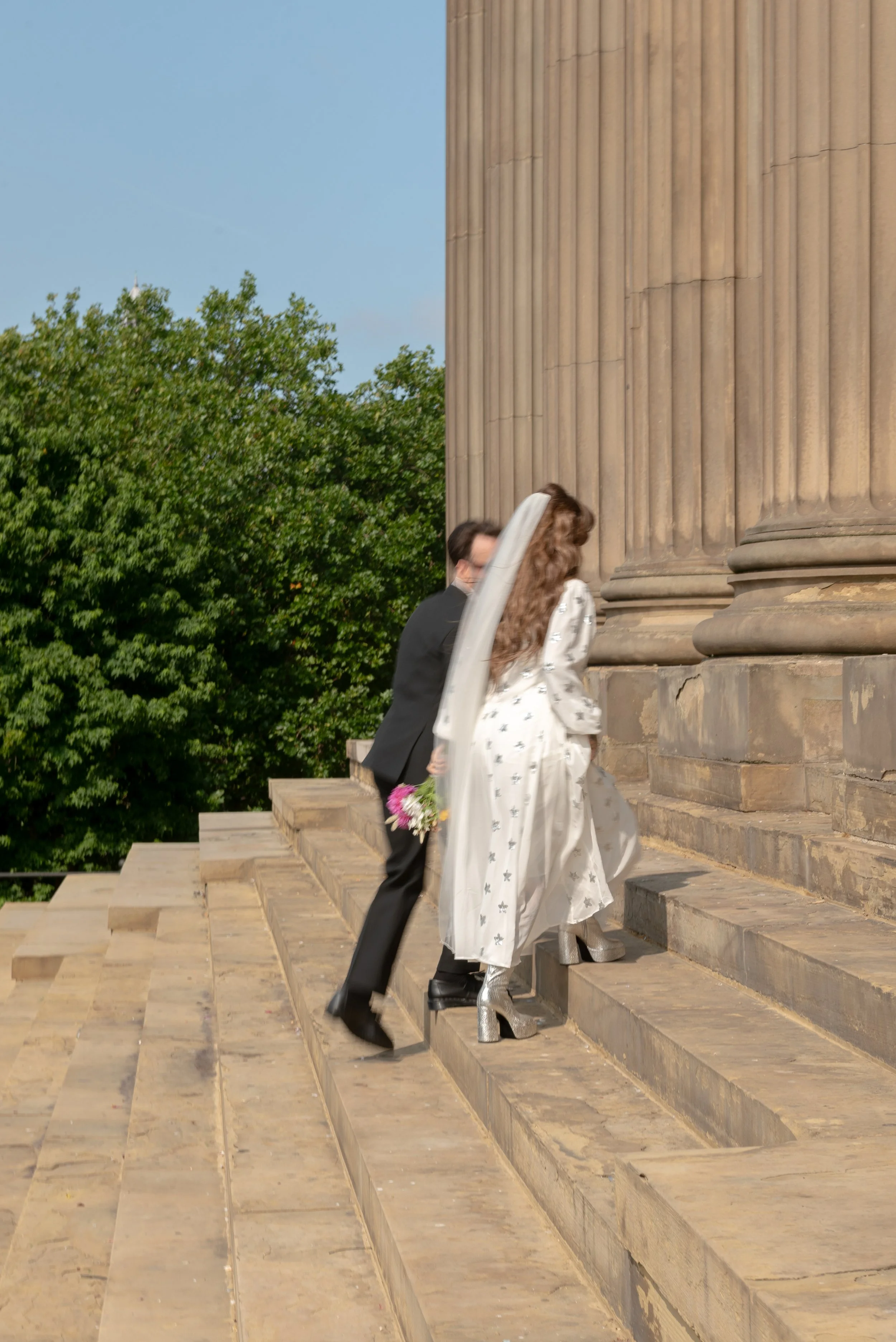 A bride in a white dress and silver heeled boots climbing the steps of a large stone building with columns, holding a bouquet of flowers, accompanied by a man in a black suit, against a backdrop of green trees.