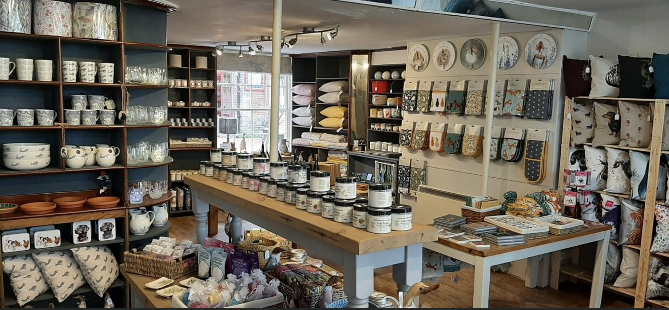 Cozy home decor store with shelves of mugs, pottery, pillows, fabric items, and wall art. Feature table displays various jars and baskets with small products.