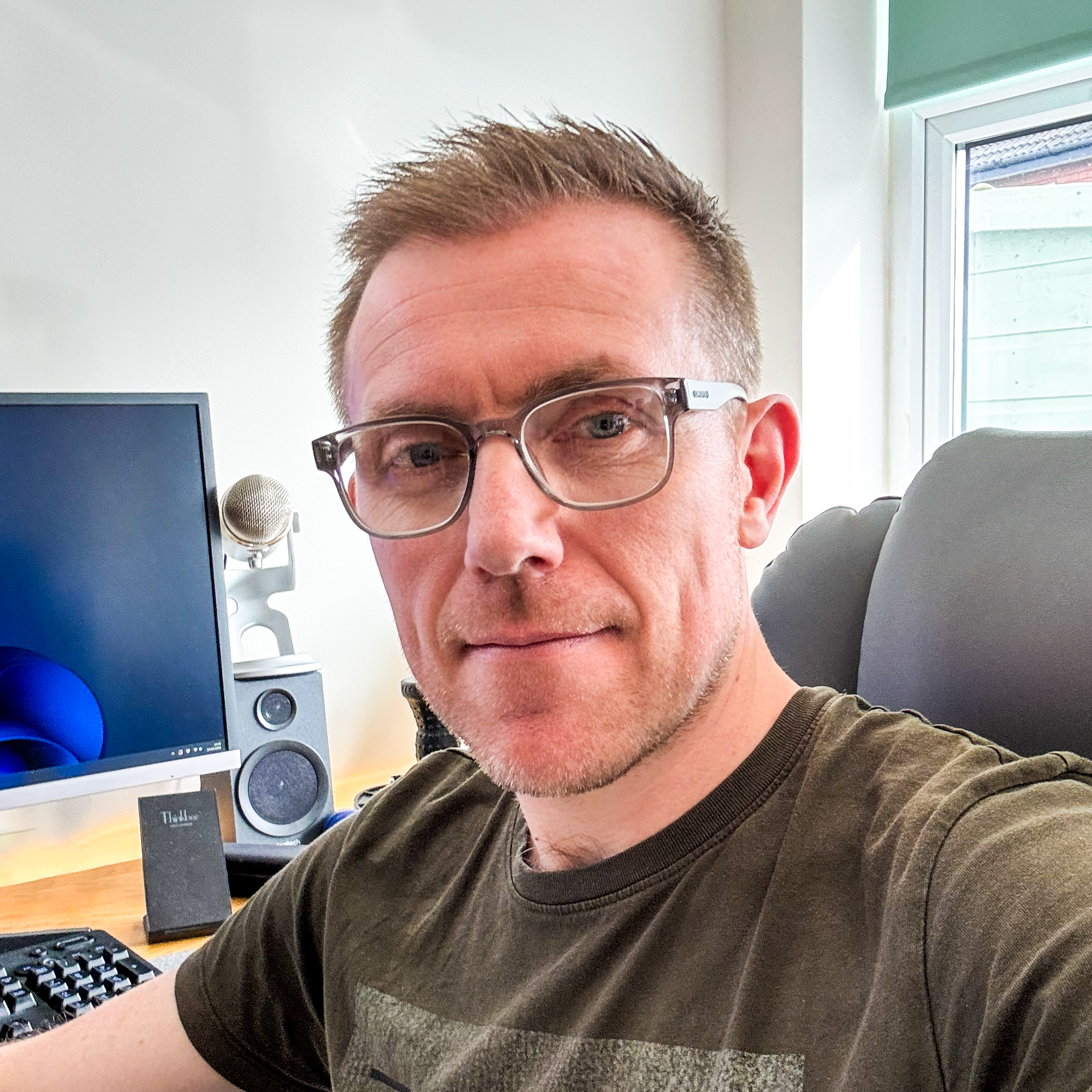 A man with glasses sitting in front of a computer with a monitor, speaker, and microphone in a bright room with a window.