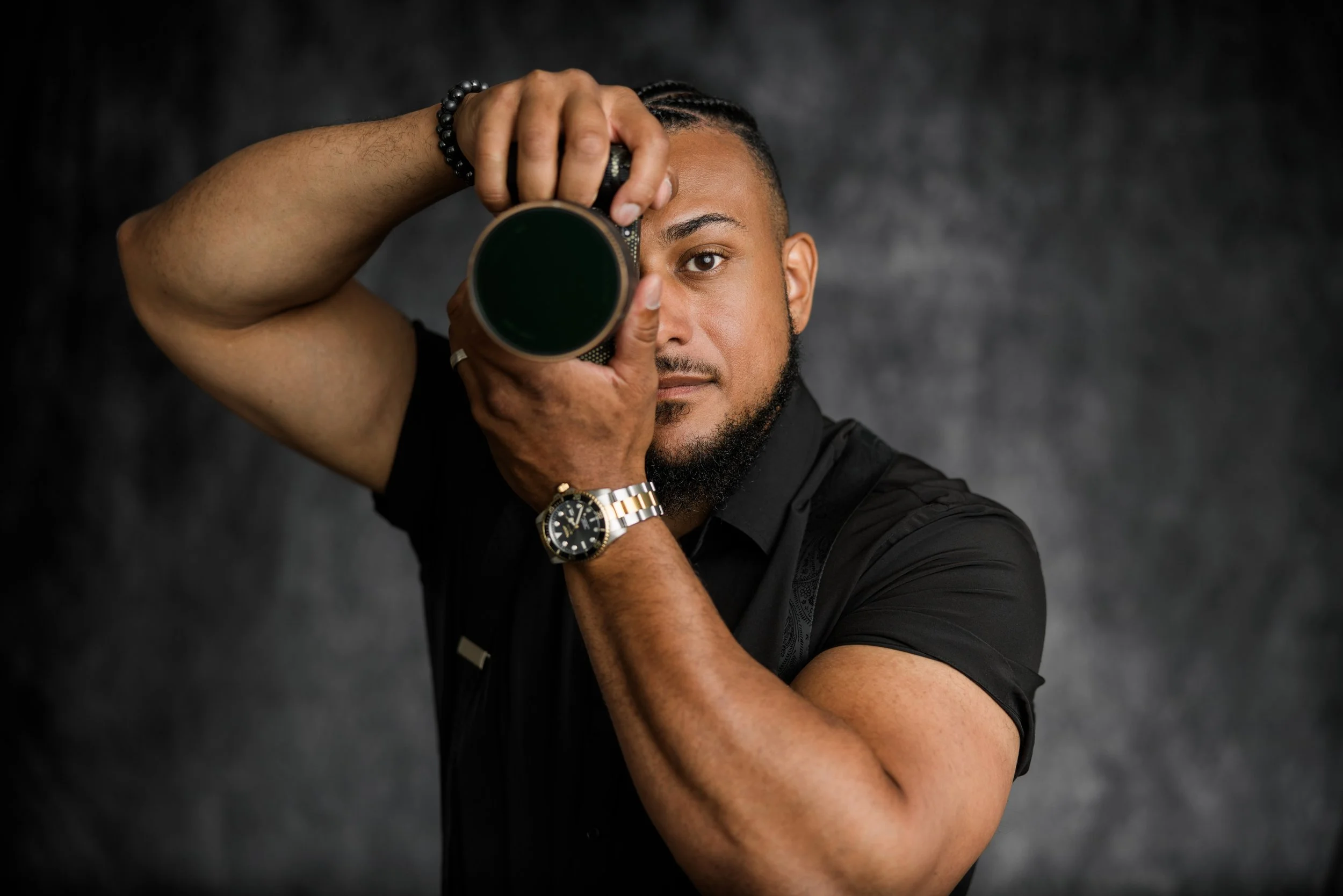 New Jersey Wedding Cinematographer Wedding Videographer with a beard and braided hair holding a camera lens in front of his eye, wearing a black shirt, a black beaded bracelet, a gold watch, and a ring on his finger, posing against a dark background.