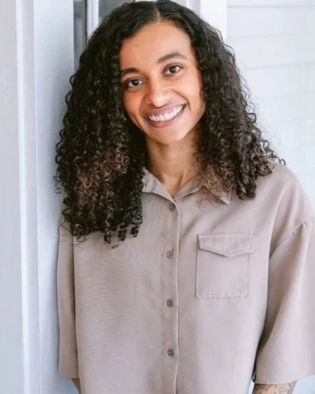 Young woman with curly dark hair smiling, wearing a beige button-up shirt and blue jeans, standing indoors near a white wall and window, with a smartwatch on her left wrist and a tattoo on her left forearm.