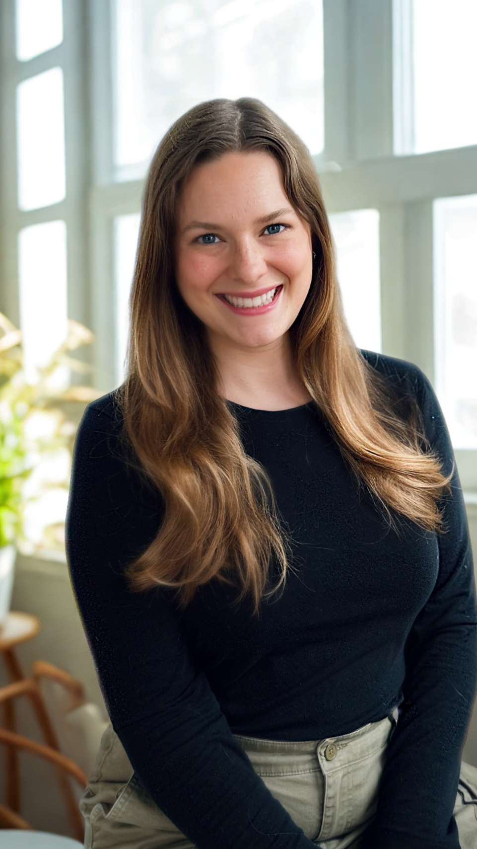 A young woman with long brown hair, blue eyes, and a bright smile, sitting on a white wicker bench with blue and white striped cushions, outdoors against a gray siding wall.