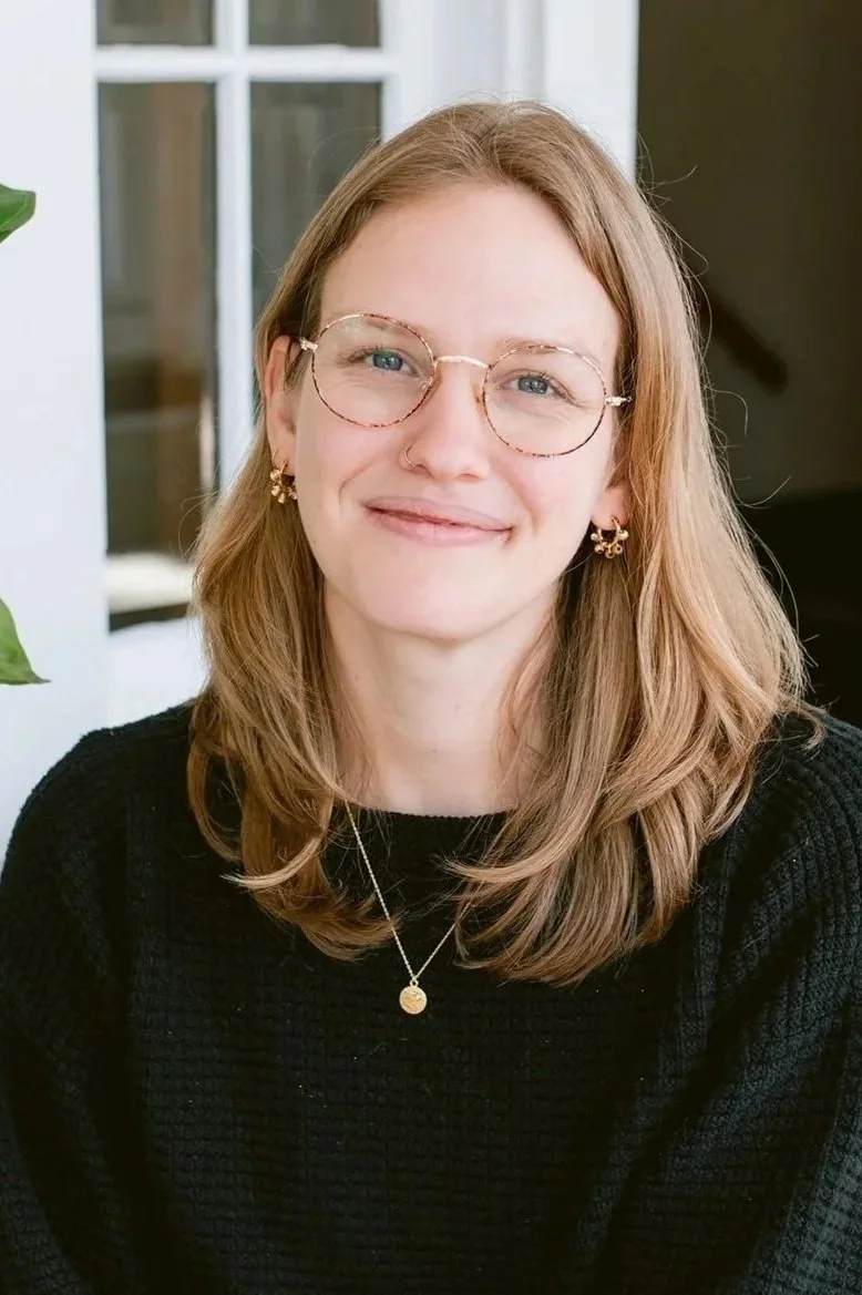 A young woman with shoulder-length light brown hair and glasses, smiling, wearing a black sweater and gold jewelry, sitting indoors near a window and a green plant.