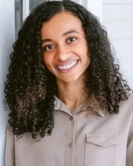 Young woman with curly dark hair smiling, wearing a beige button-up shirt and blue jeans, standing indoors near a white wall and window, with a smartwatch on her left wrist and a tattoo on her left forearm.