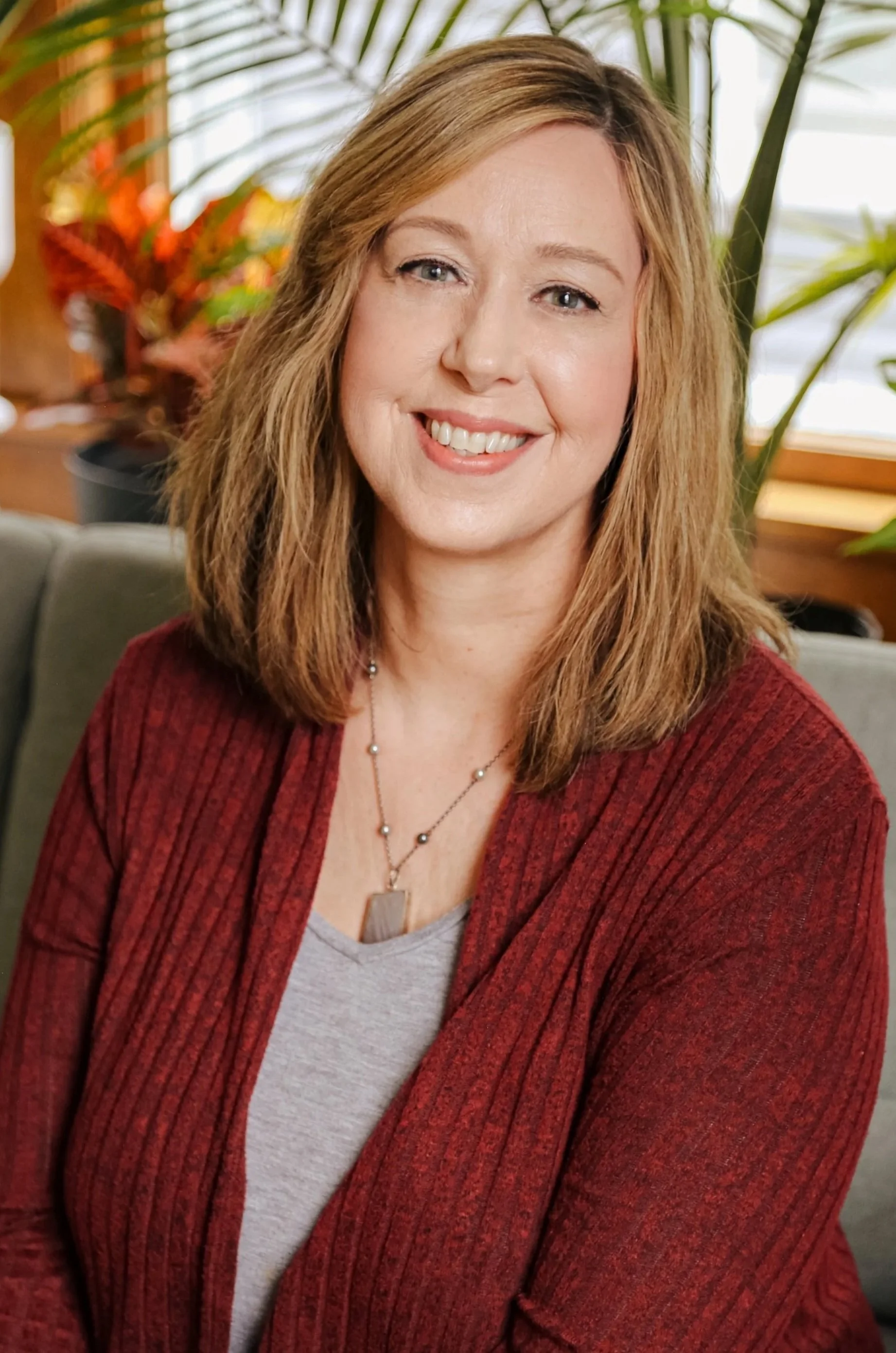 A woman with shoulder-length light brown hair, wearing a gray top and a red cardigan, smiling while sitting on a gray couch in a room with green plants and wooden window frames.