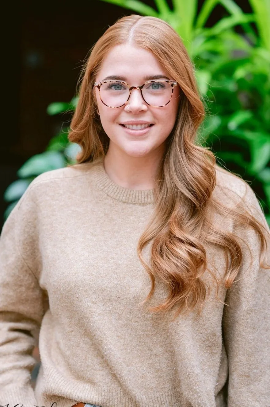 Portrait of a young woman with long red hair, light skin, wearing glasses, a beige sweater, and jeans, standing outdoors with green plants in the background.