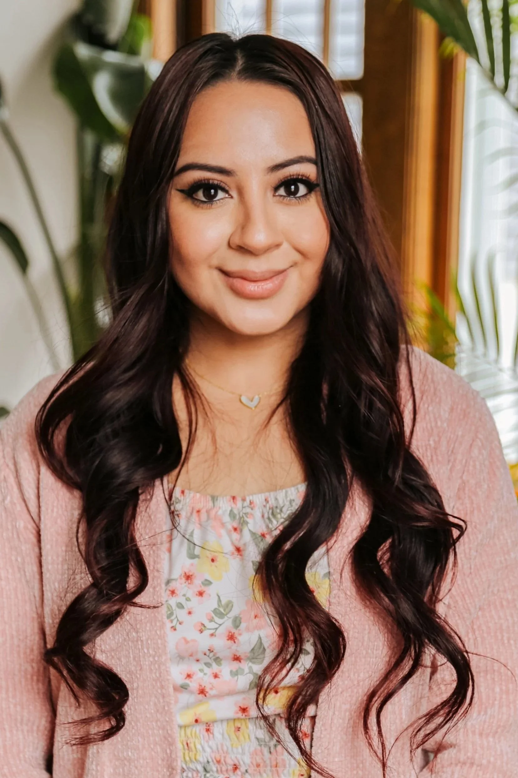 Mental health therapist who speaks Spanish and English. A young woman with long, dark, wavy hair, smiling, wearing a pink cardigan, floral top, and blue button-up pants, standing indoors with green plants and a window in the background.