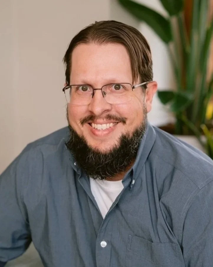 A smiling man with glasses and a beard wearing a blue button-up shirt, seated indoors near a plant.