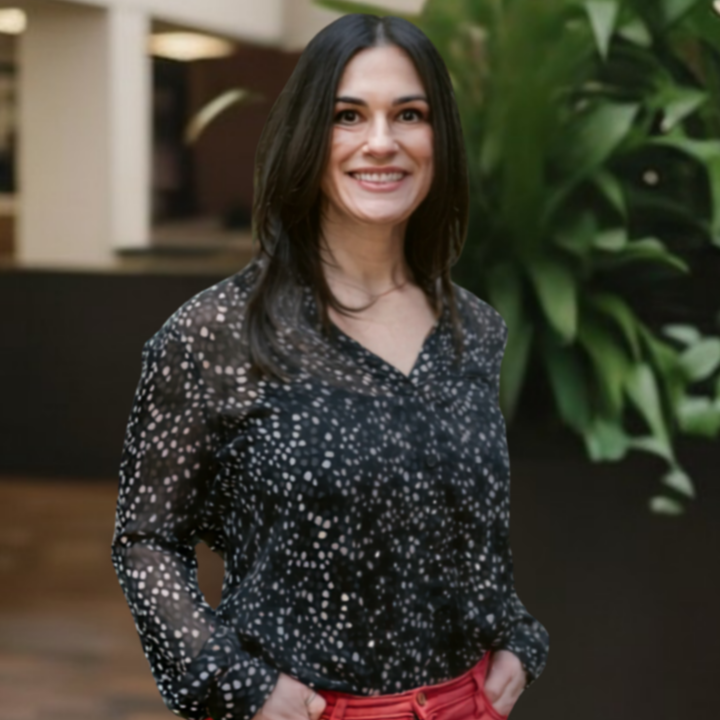 A woman with long dark hair, smiling, wearing a black and white patterned blouse, standing in front of green indoor plants.