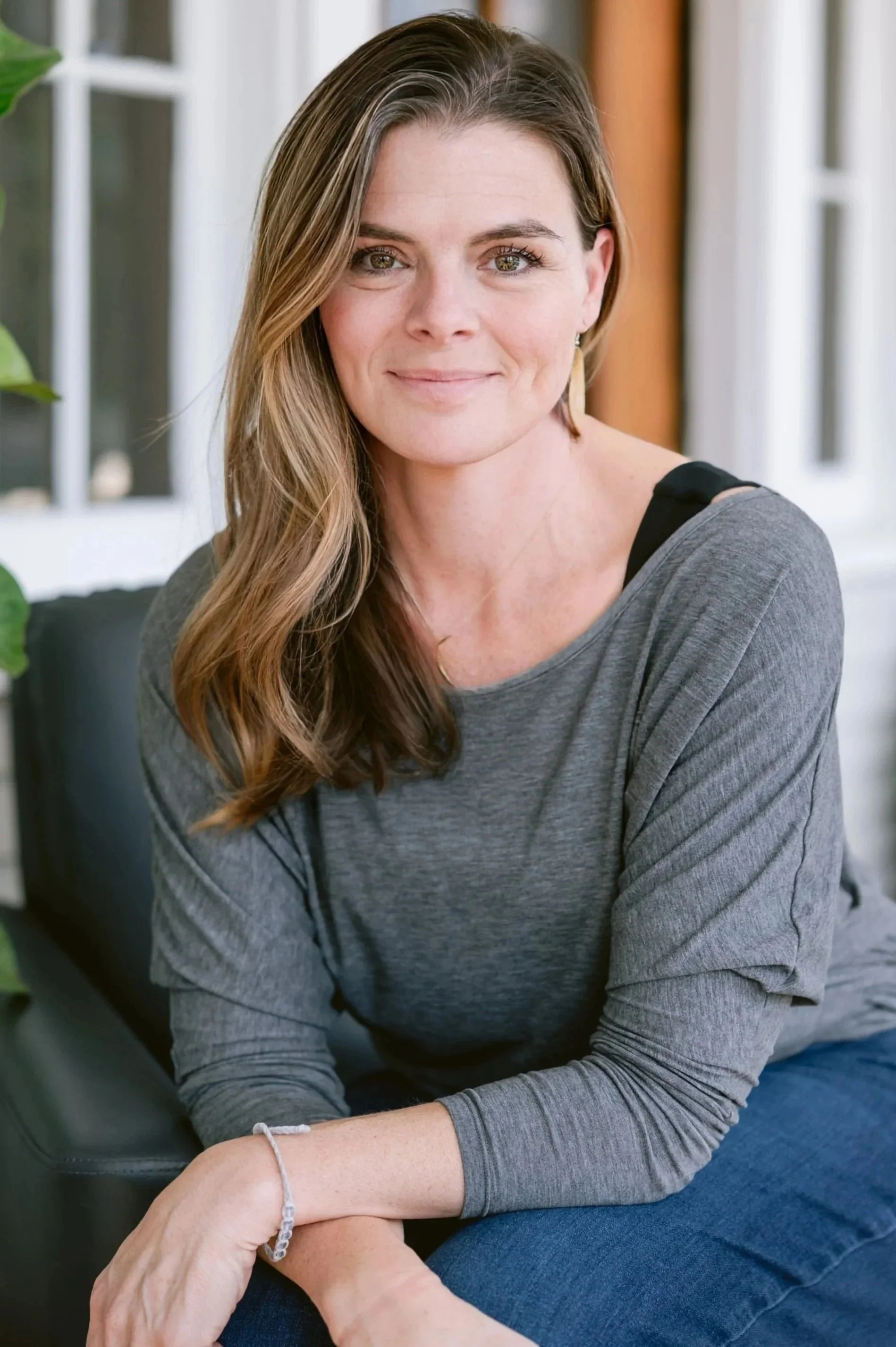 A woman with long wavy brown hair, wearing a gray long-sleeve shirt and blue jeans, sitting on a black chair on a porch with white and wood siding, surrounded by potted plants and greenery.