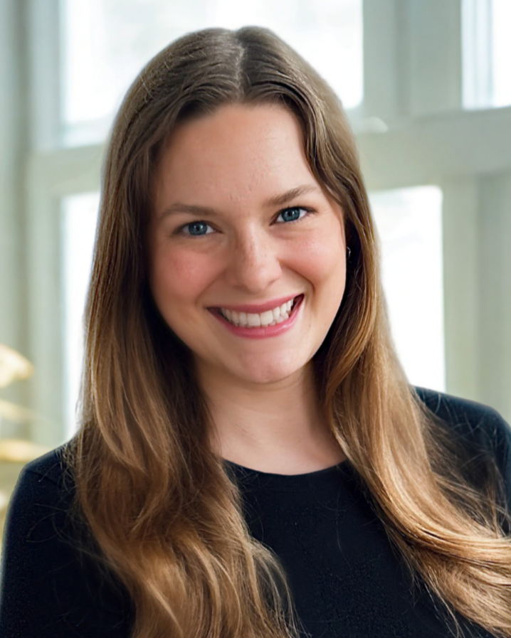 A young woman with long brown hair, blue eyes, and a bright smile, sitting on a white wicker bench with blue and white striped cushions, outdoors against a gray siding wall.