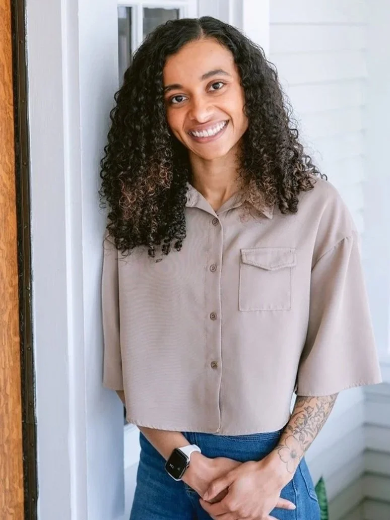 Young woman with curly dark hair smiling, wearing a beige button-up shirt and blue jeans, standing indoors near a white wall and window, with a smartwatch on her left wrist and a tattoo on her left forearm.
