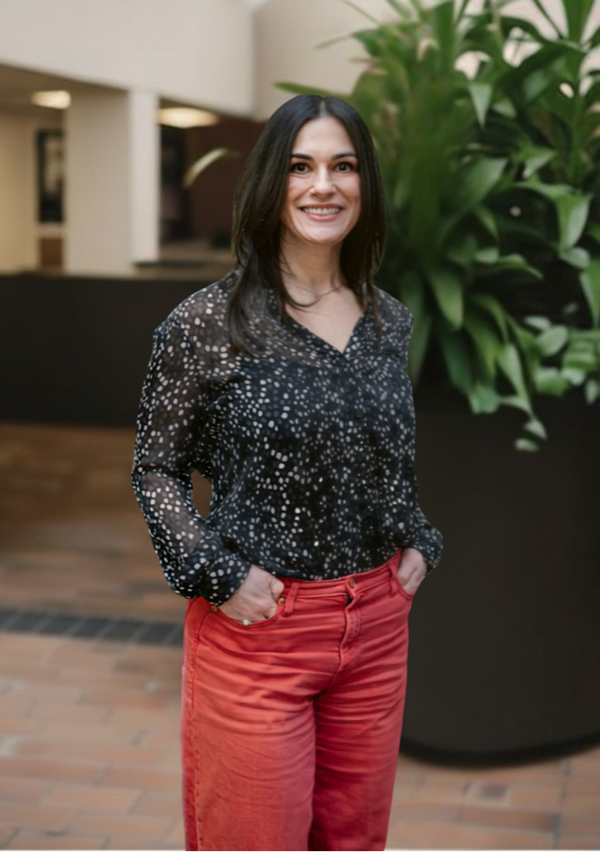 A woman with long dark hair, smiling, wearing a black and white patterned blouse, standing in front of green indoor plants.