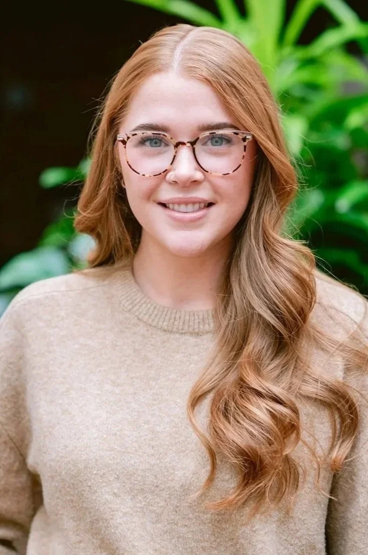 Portrait of a young woman with long red hair, light skin, wearing glasses, a beige sweater, and jeans, standing outdoors with green plants in the background.