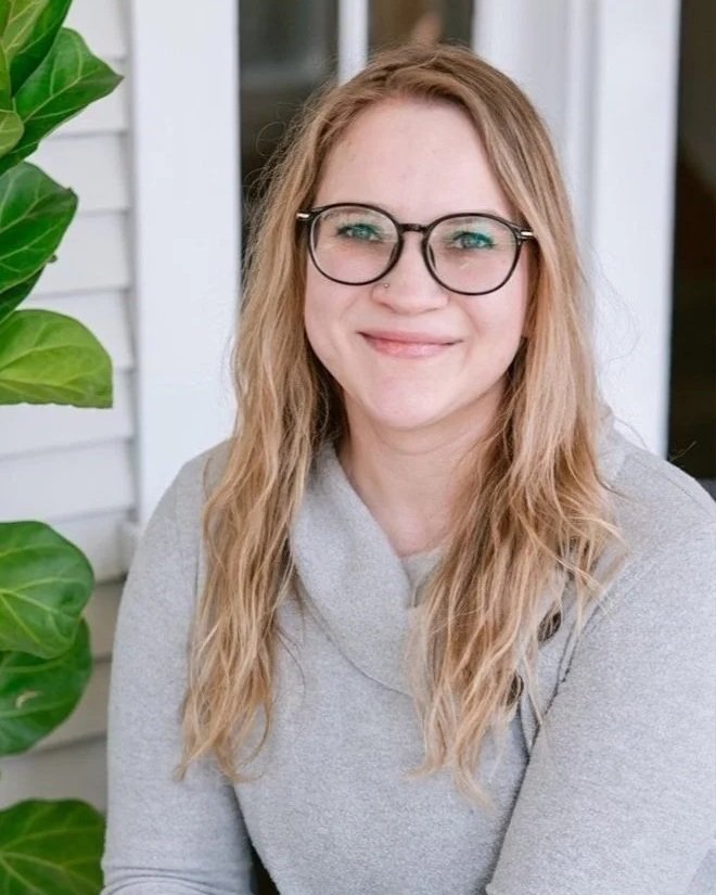 A smiling woman with long wavy blonde hair and glasses sitting outdoors near a house with white siding and large green leafy plants.