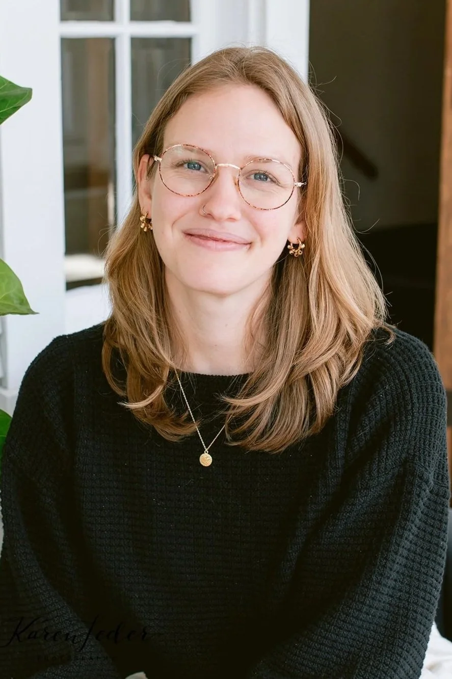 A young woman with shoulder-length light brown hair and glasses, smiling, wearing a black sweater and gold jewelry, sitting indoors near a window and a green plant.