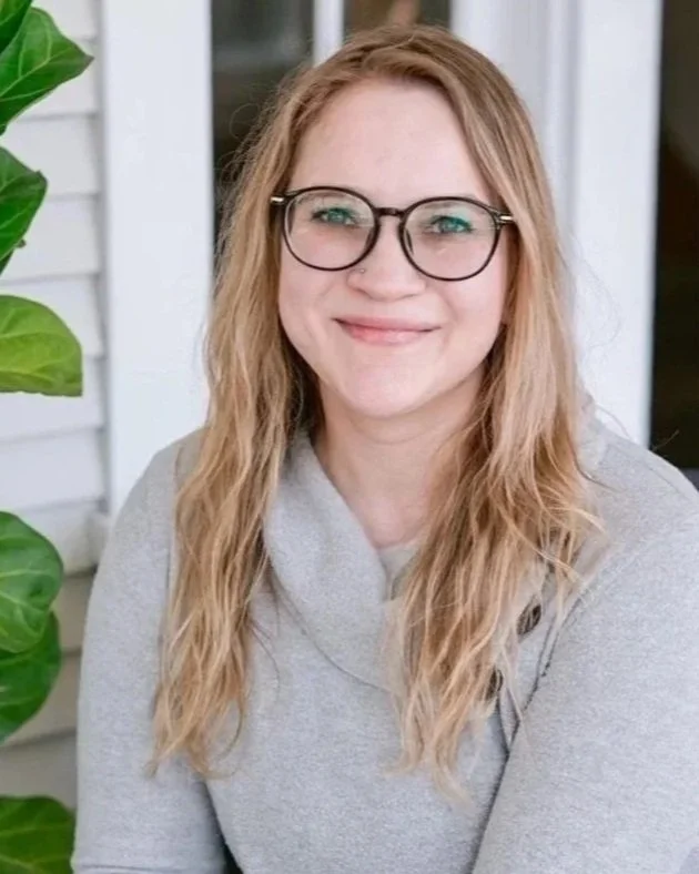 A smiling woman with long wavy blonde hair and glasses sitting outdoors near a house with white siding and large green leafy plants.