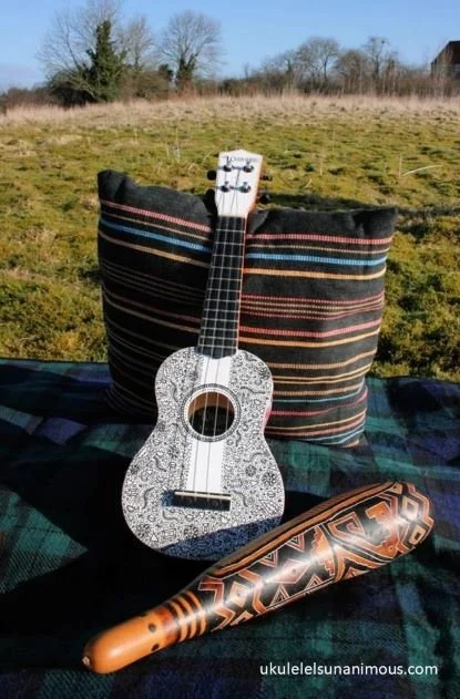 White ukulele in a field on a picnic rug with cushion.