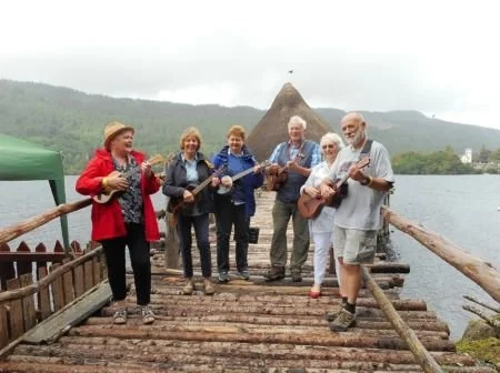 Group of people playing ukuleles by a lake.