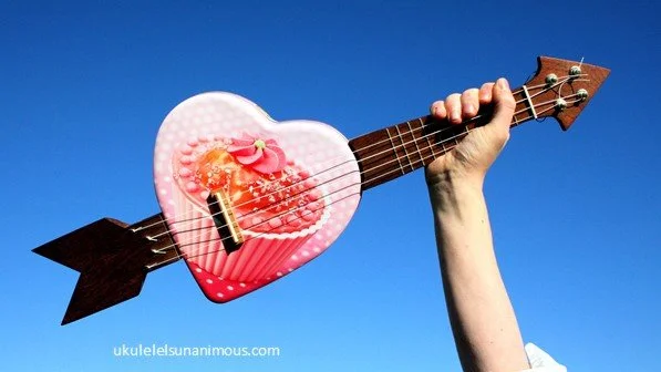 Pink love heart cup cake ukulele held up against a blue sky.
