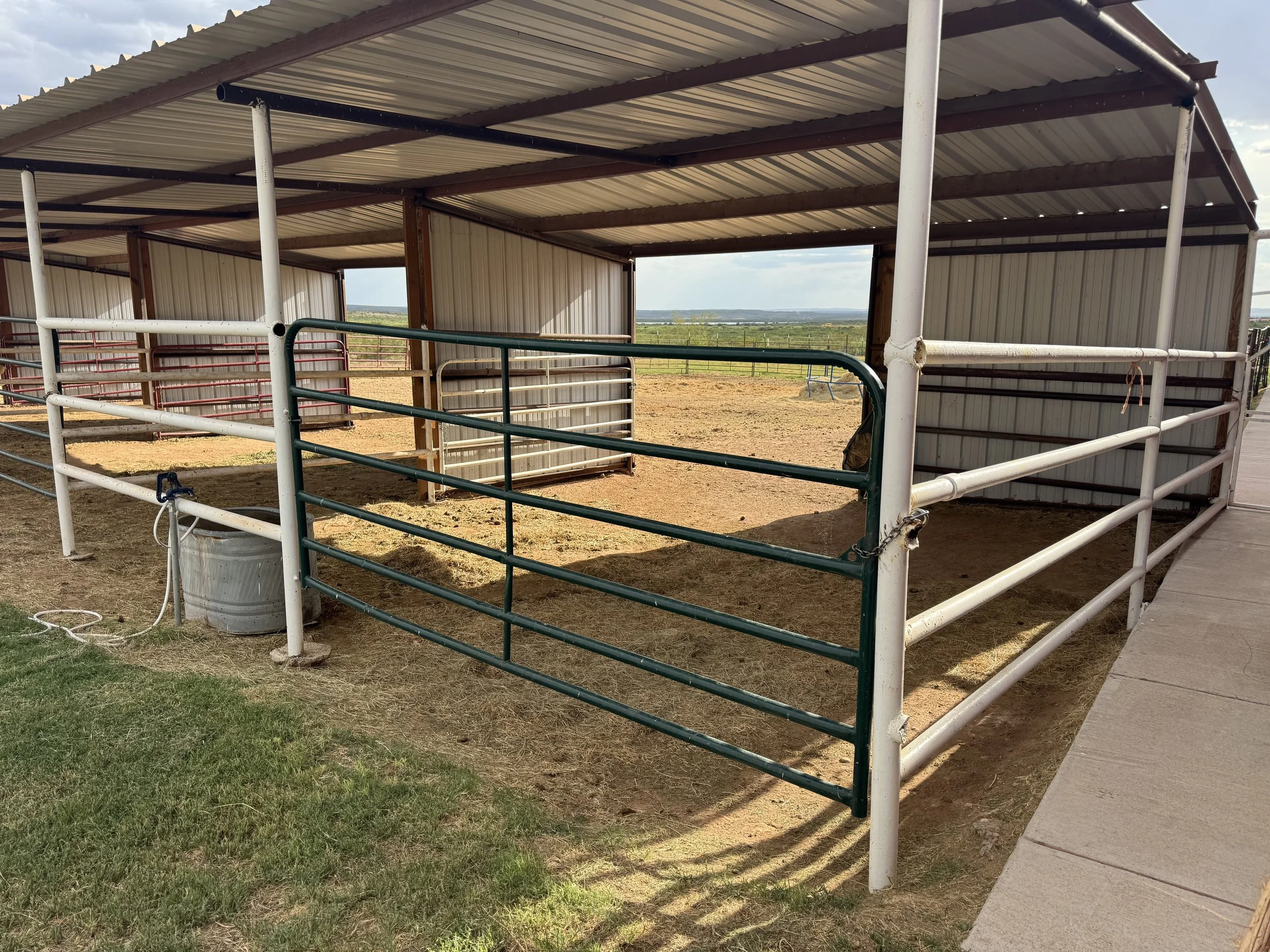 Open animal enclosure or barn with metal and wood fencing, metal roof, and view of open countryside in the background.