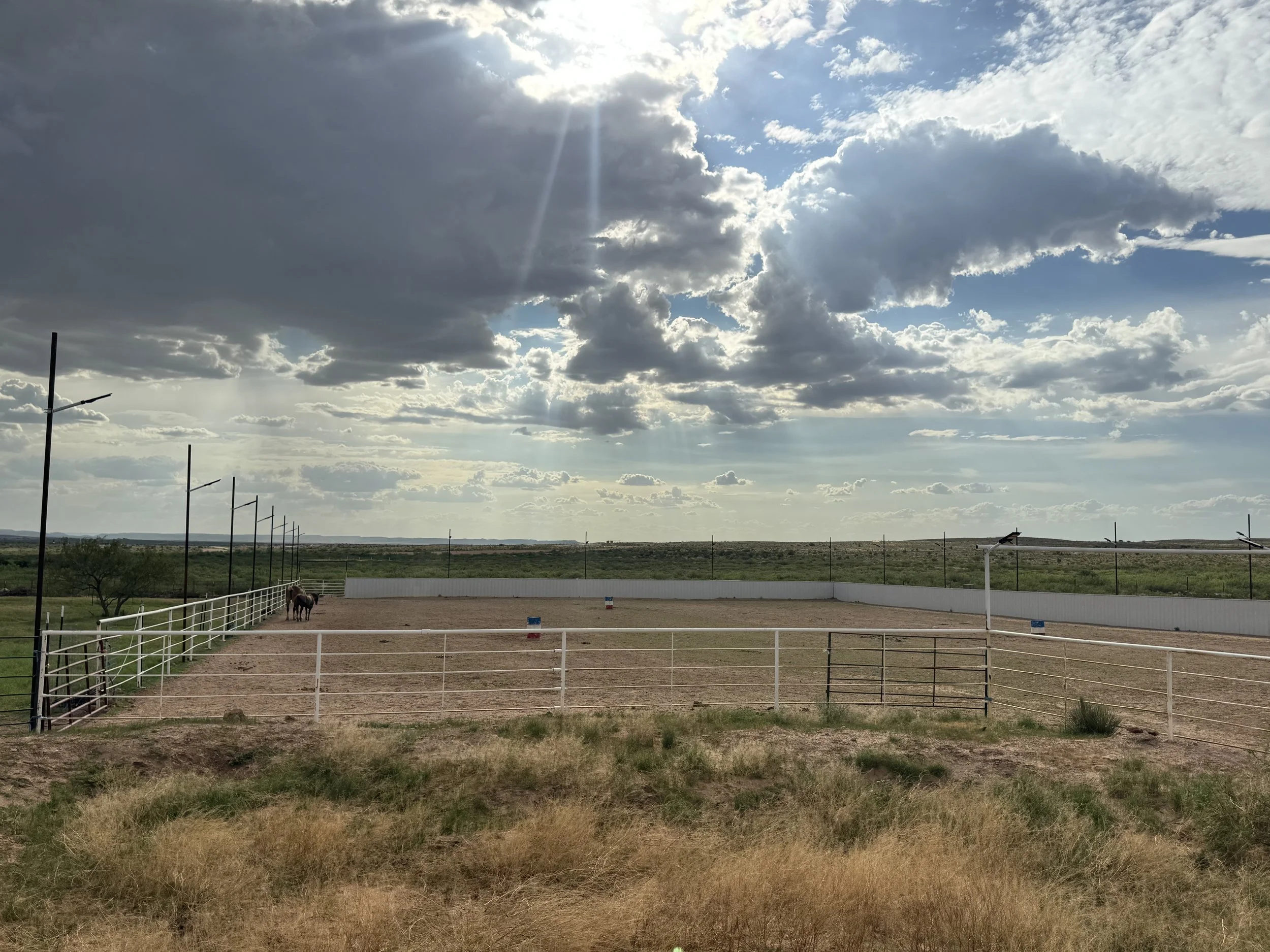 A large outdoor horse riding arena with a horse near the fence, under a partly cloudy sky with sun rays breaking through.