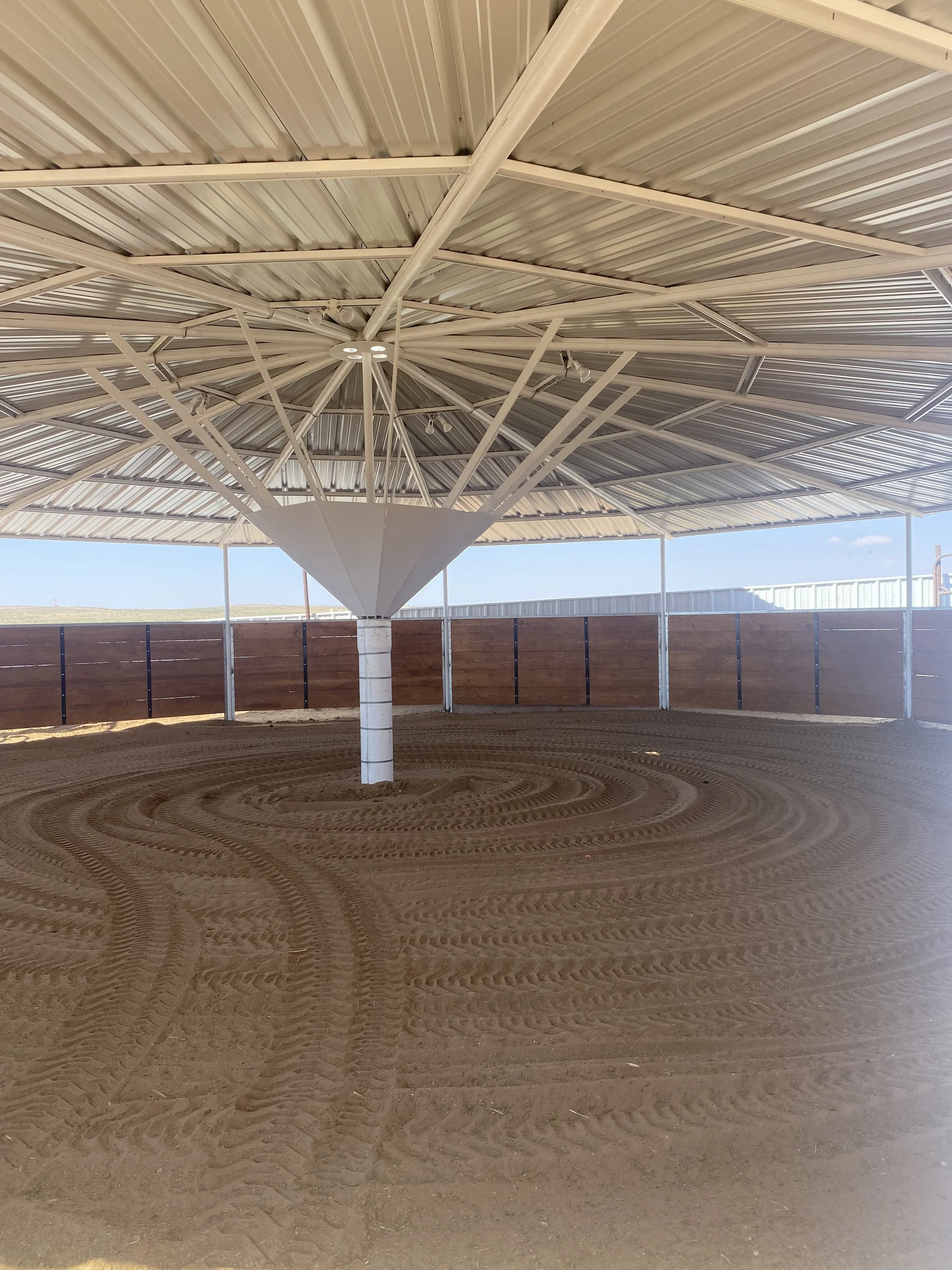 A large, open-air structure with a metal roof supported by a central pole and beams, with a circular, patterned dirt floor underneath.