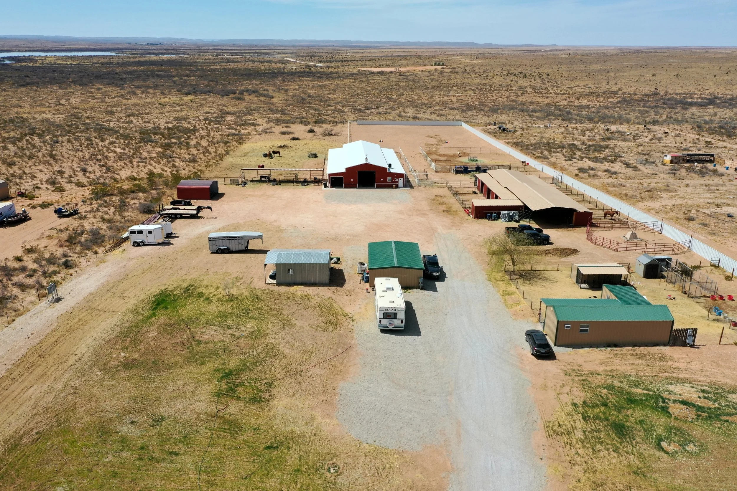 An aerial view of a rural property with a large red barn, surrounded by open land and sparse desert vegetation, with several parked vehicles and trailers.