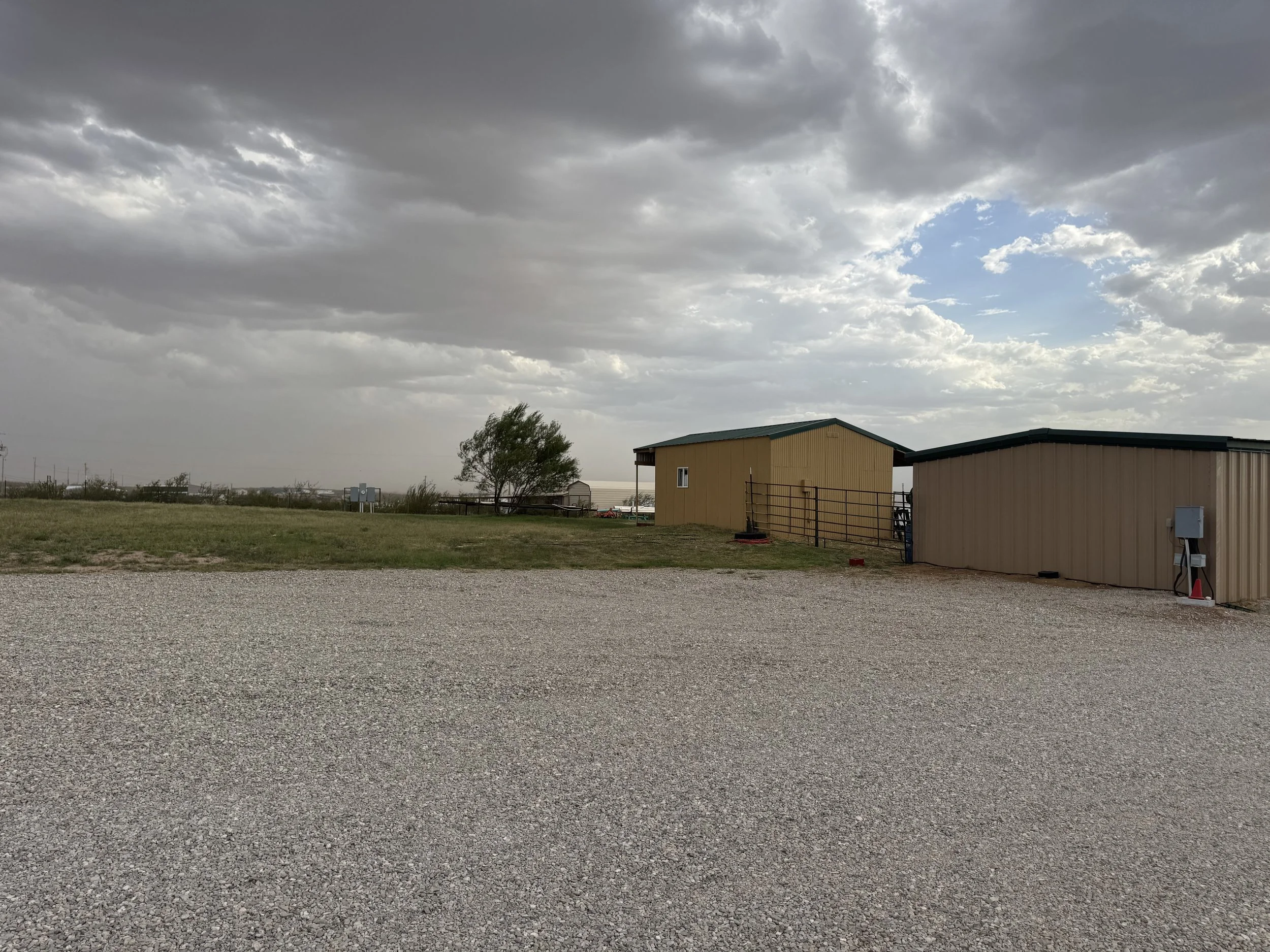 A gravel parking lot in front of two beige buildings with green roofs, a tree, and overcast sky with dark clouds and patches of blue.