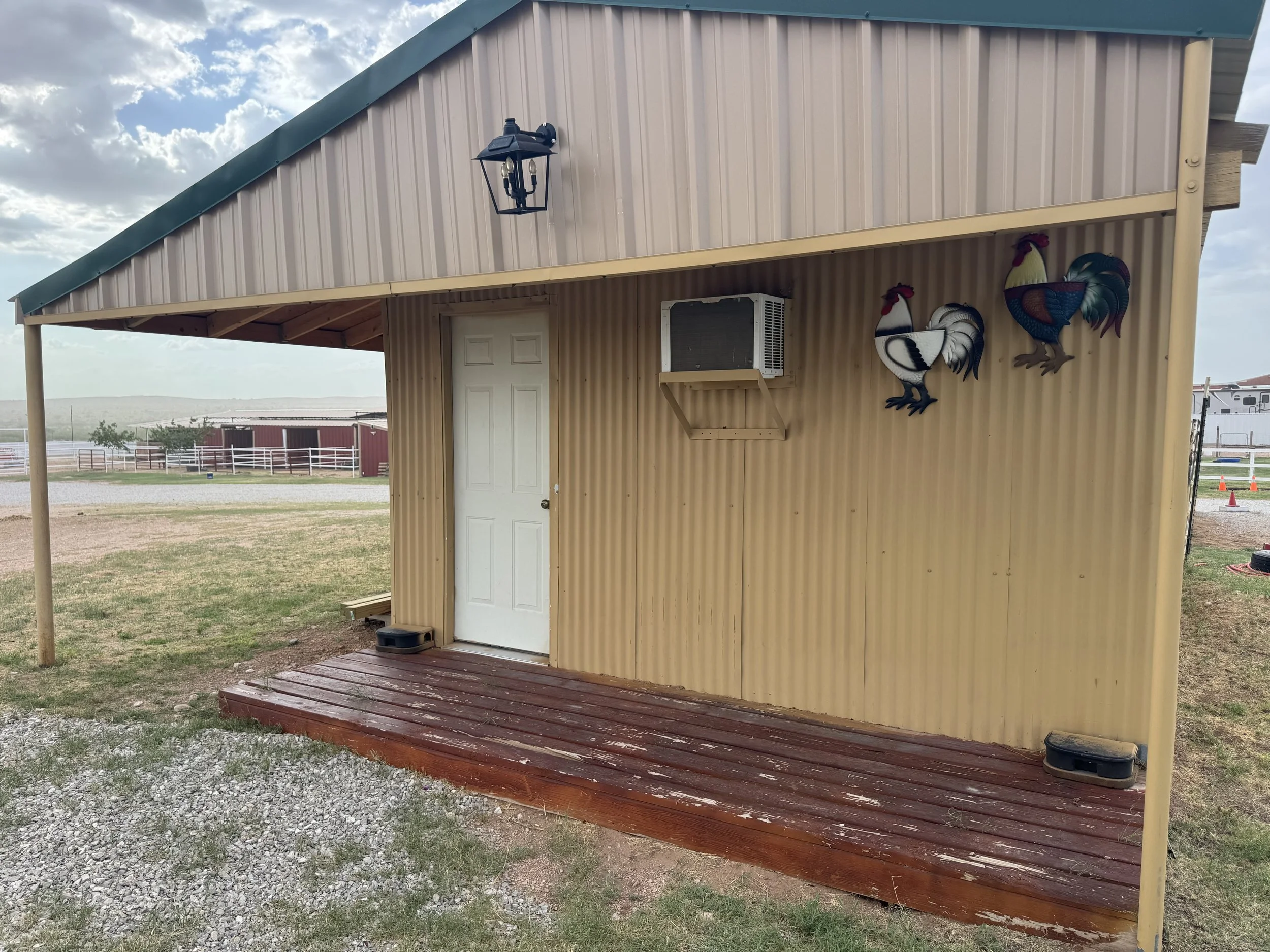A small beige metal building with a white door, a mounted window air conditioning unit, decorative rooster wall art, and a black outdoor lantern light fixture. The building is on a wooden deck with gravel and grass in the foreground and open field and sky in the background.