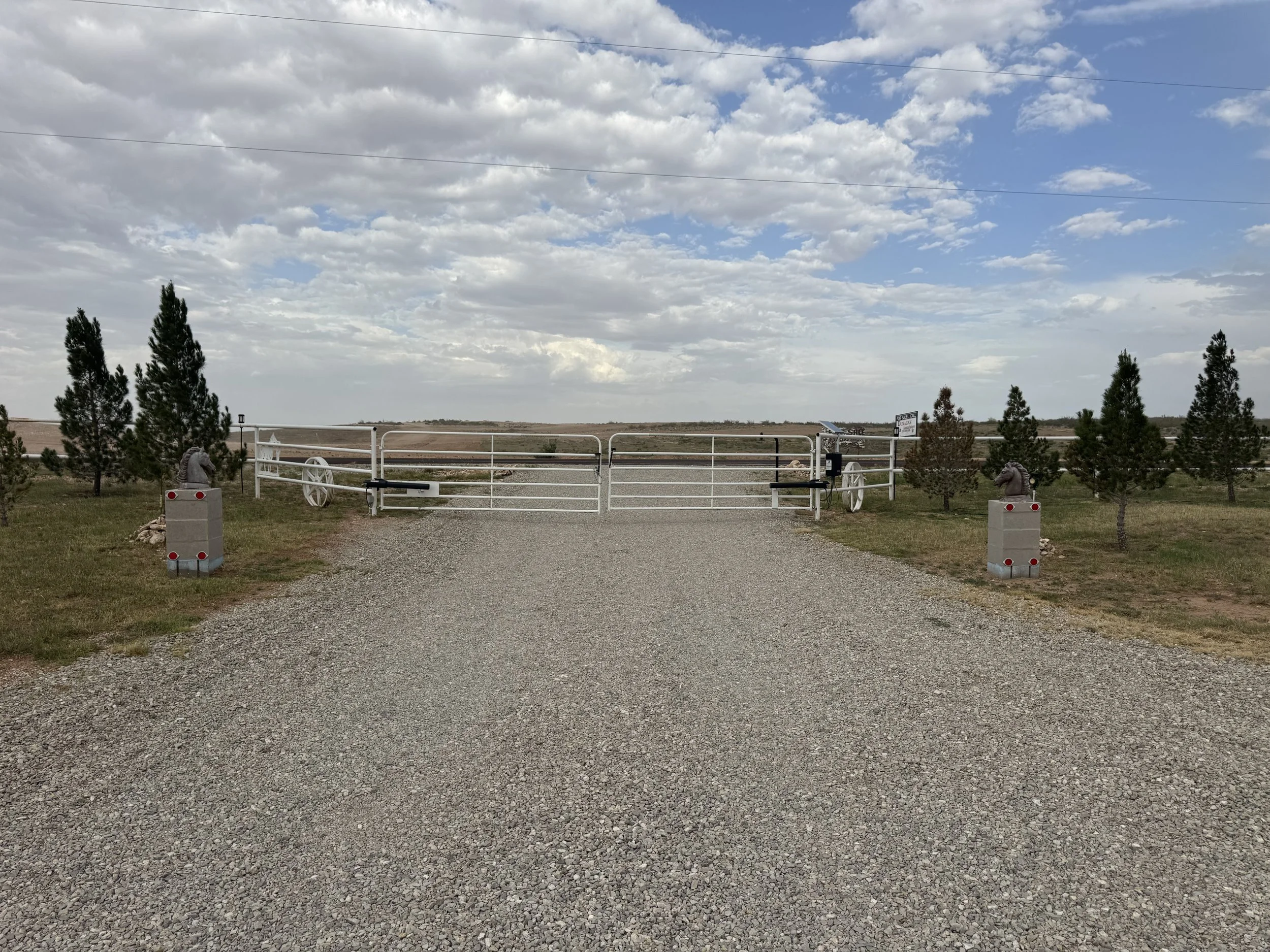 A gravel driveway leading to a white metal gate with trees on either side, and a landscape of open land and a cloudy sky in the background.