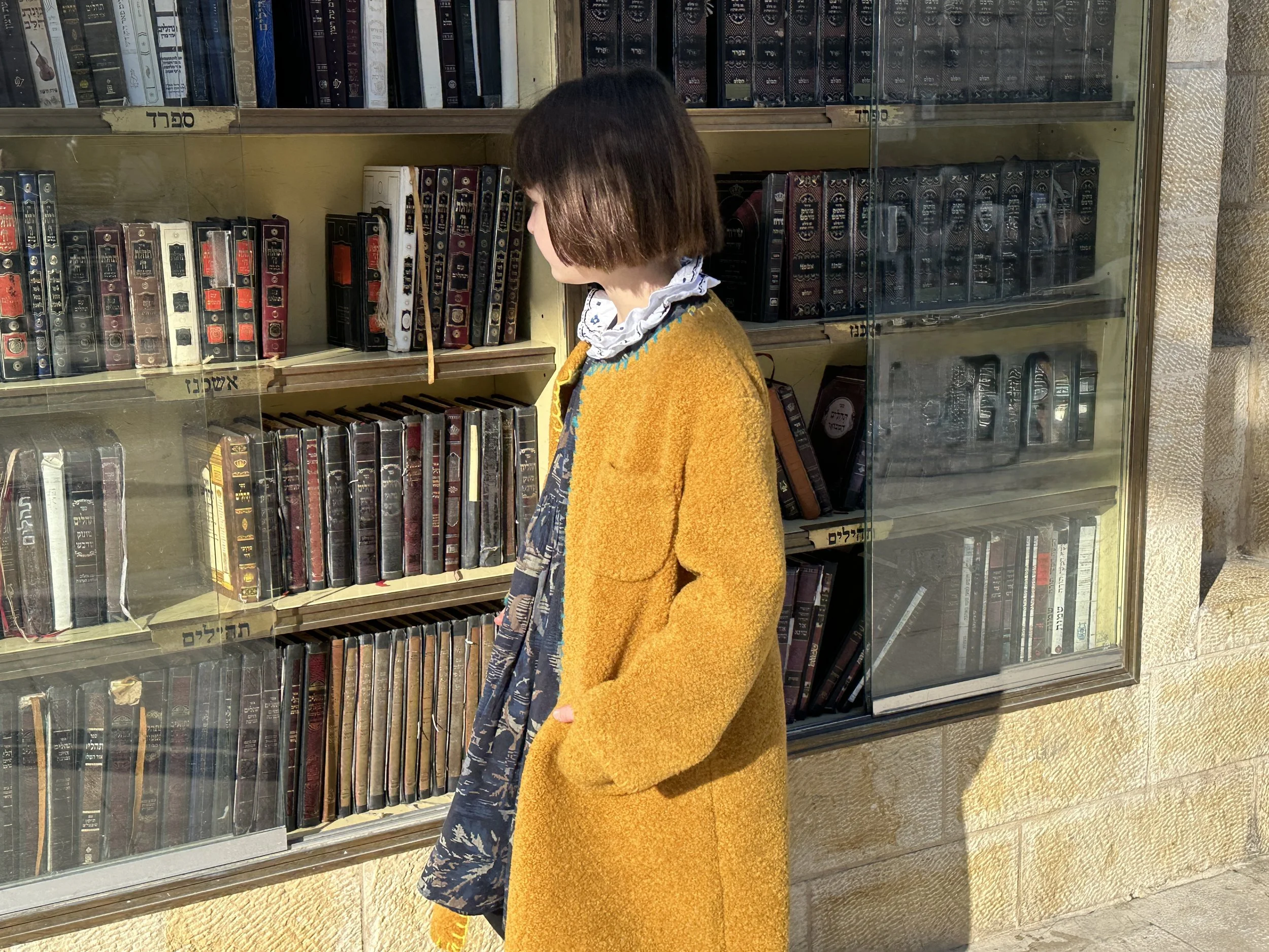 A woman wearing a yellow coat and a patterned scarf stands outside a bookstore, looking at a glass display window filled with books.