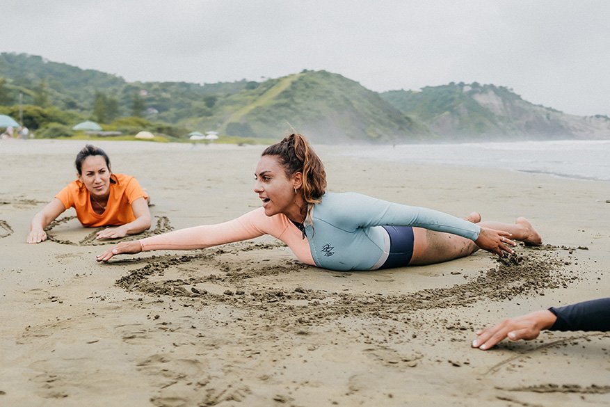 Tres mujeres en la playa practicando yoga en arena húmeda, una de ellas extendiendo los brazos y cuerpo en posición de largo en el suelo, mientras las otras dos observan.