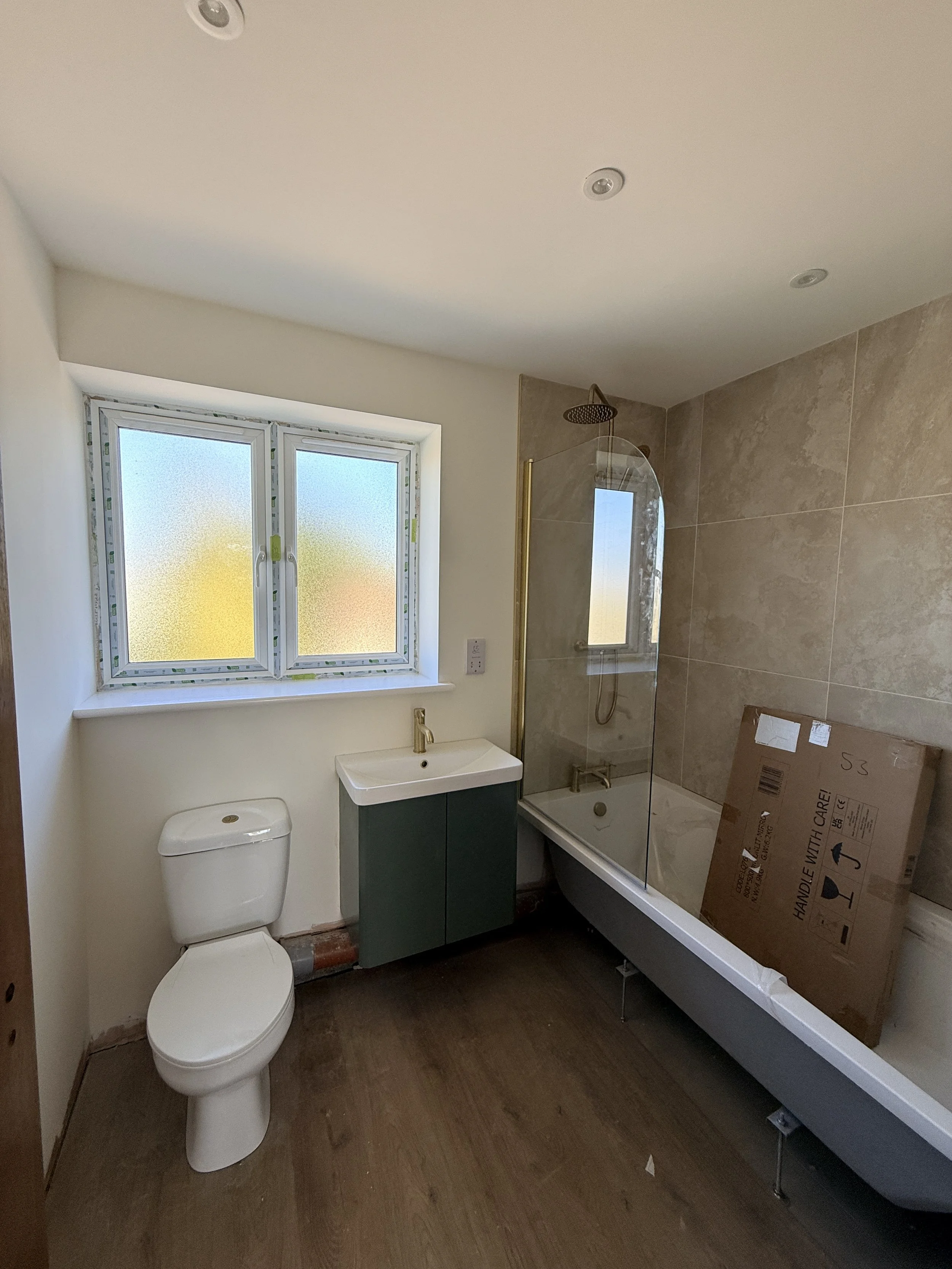 A newly renovated bathroom with a toilet, a small green vanity with a sink, and a walk-in shower with a glass screen and a cardboard box inside.