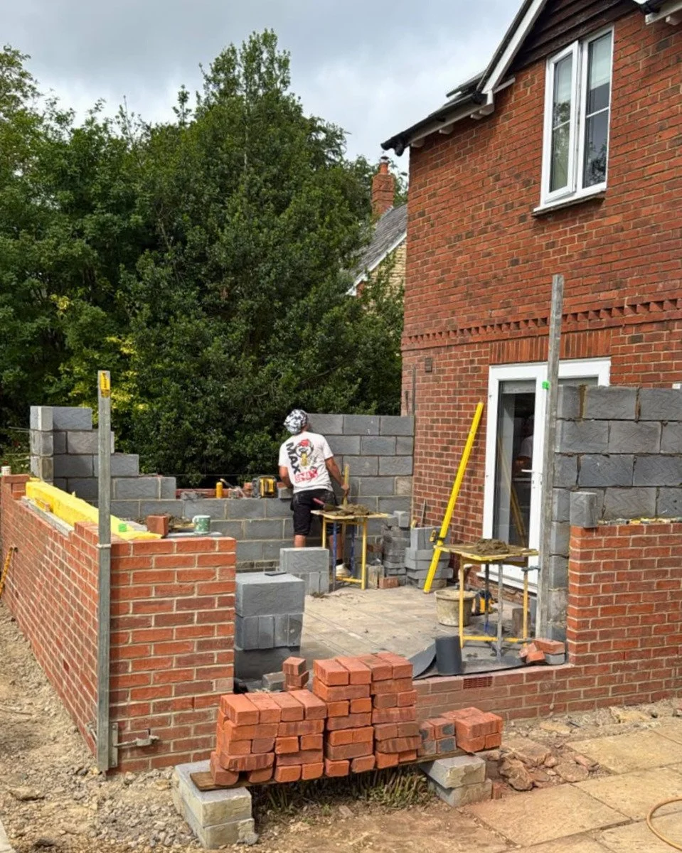 A person is building a brick wall outside a red brick house. The construction site has bricks, tools, and scaffolding around. The person is working on the wall, which is partially built.