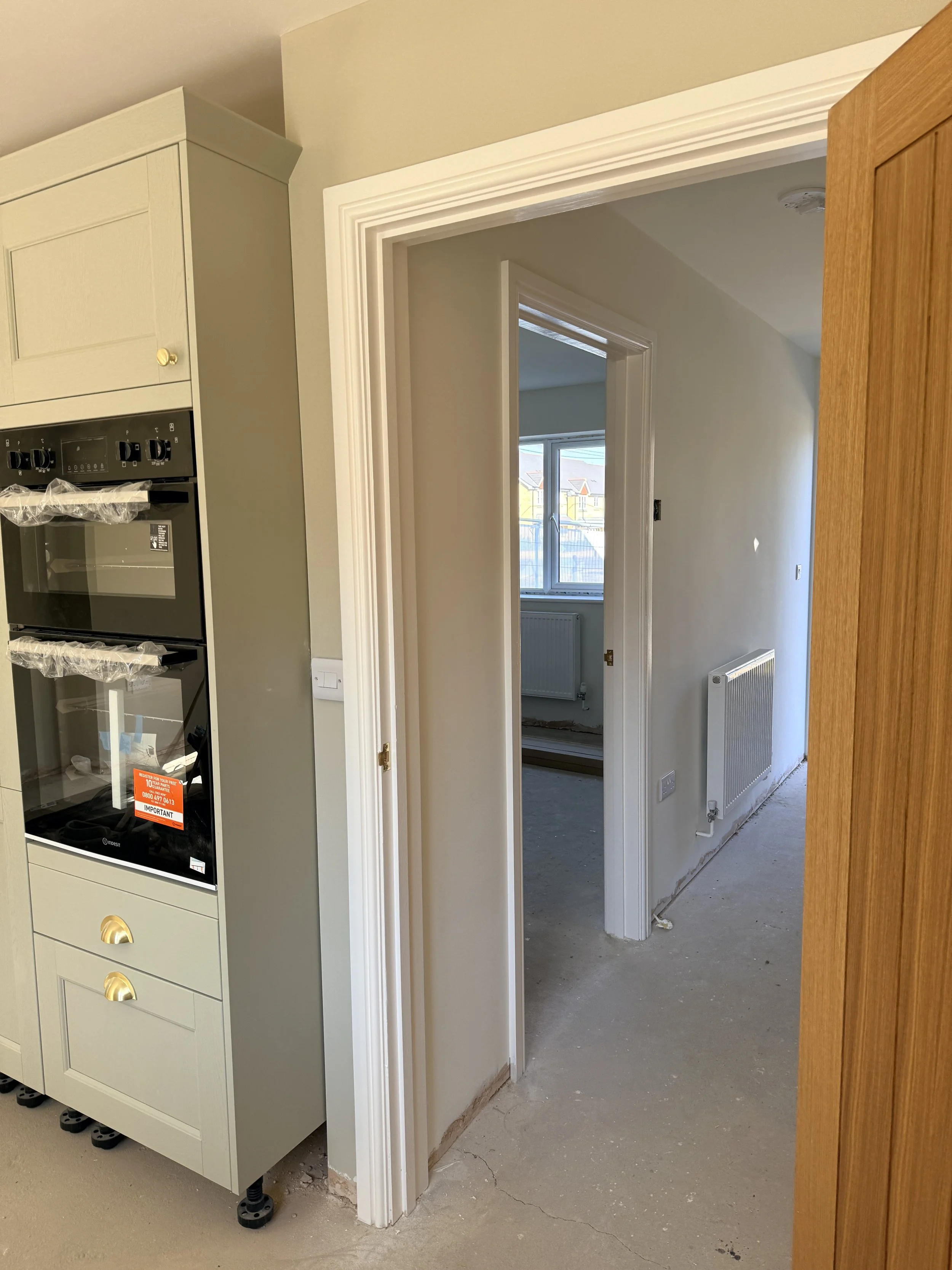 A view of an interior hallway with a doorway showing another room, a radiator, and part of kitchen cabinetry including an oven, indicating a home under renovation.