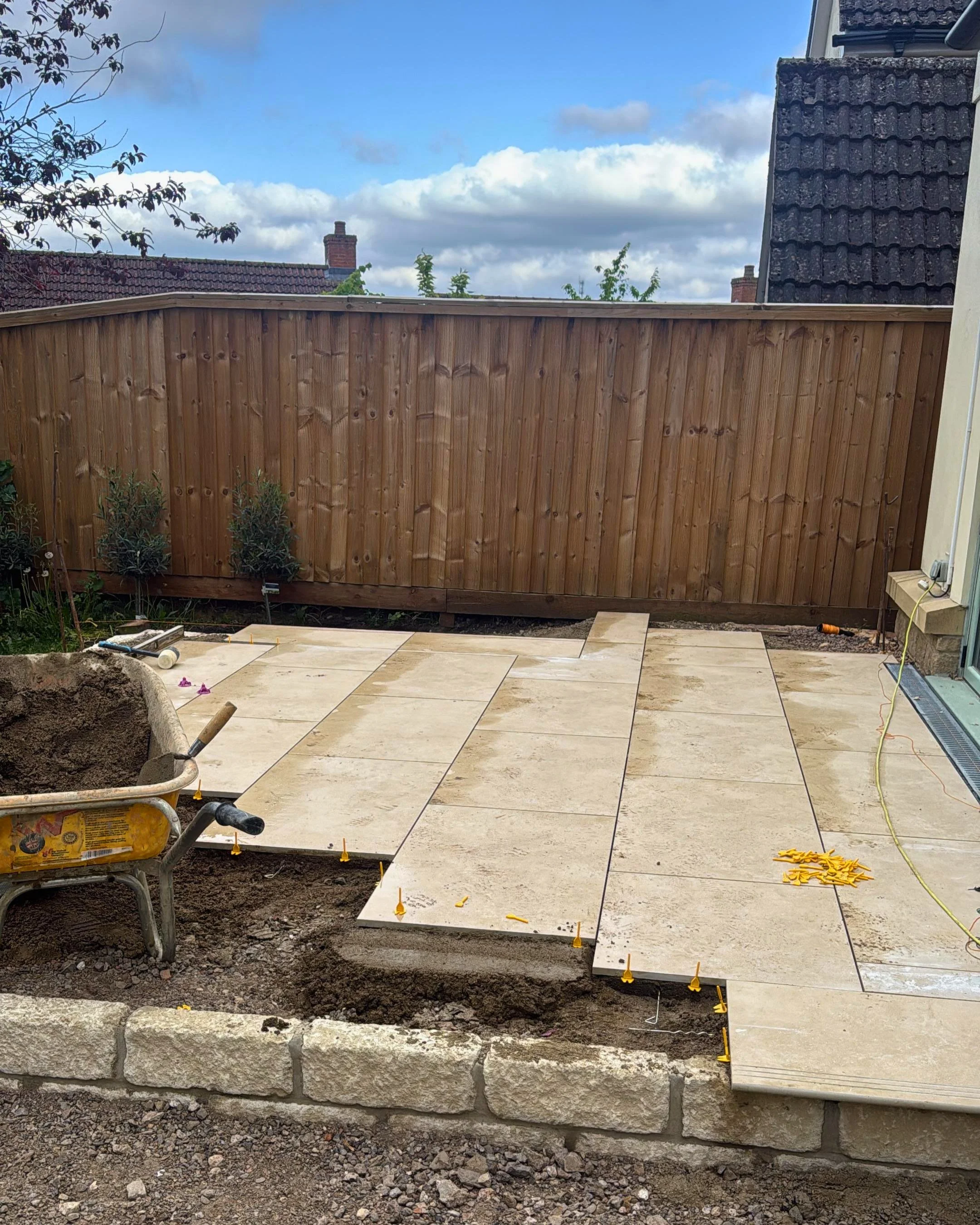 Backyard construction site with new stone pavers being installed, a wooden privacy fence, and construction tools and materials visible, with a house and cloudy sky in the background.