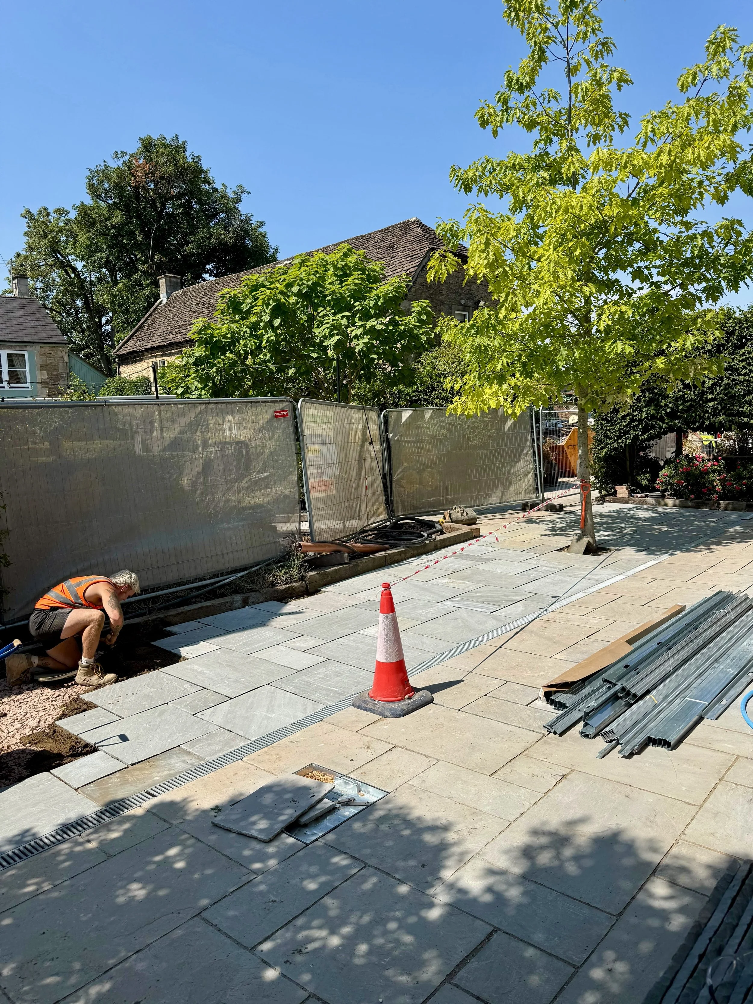 A construction worker installing gray paving stones on a walkway next to a tree, with metal pipes stacked nearby and a traffic cone marking the work area, in a residential neighborhood under a clear blue sky.