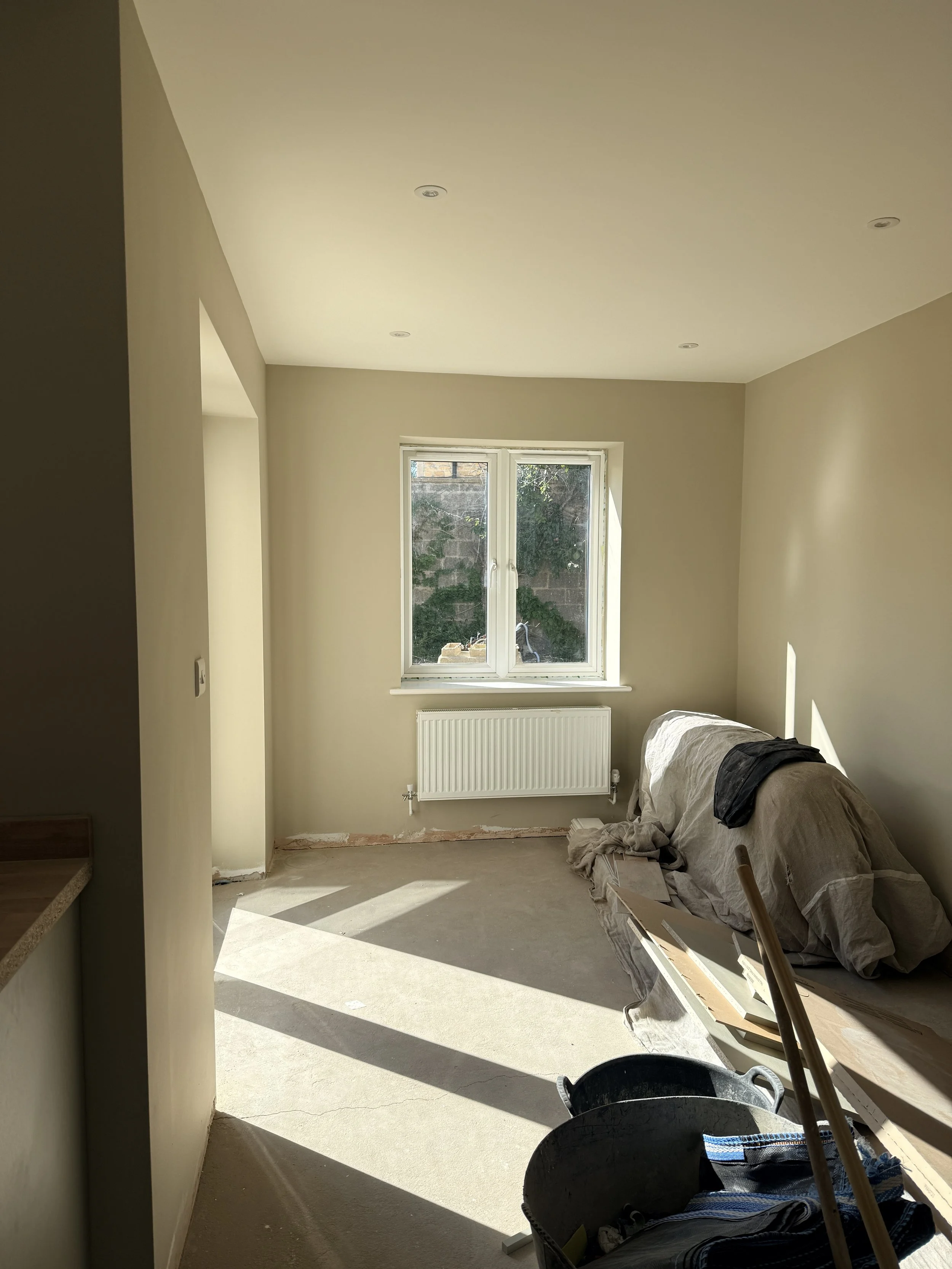 A room under construction with a window, a white radiator, and construction materials including a black bucket and tools.