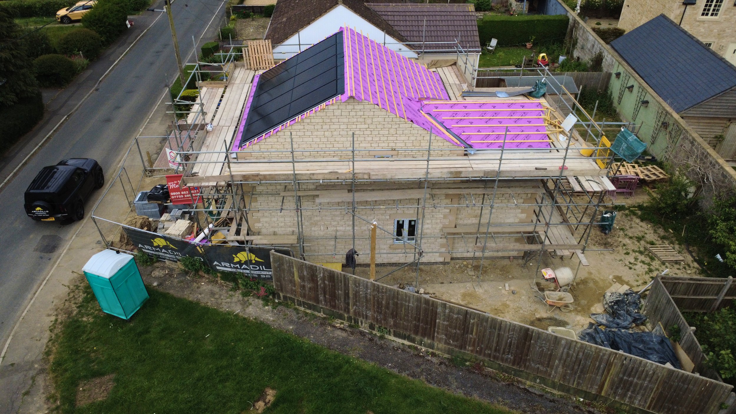 A house under construction with scaffolding around it, showing half-completed roofwork and black roof tiles on the finished section.
